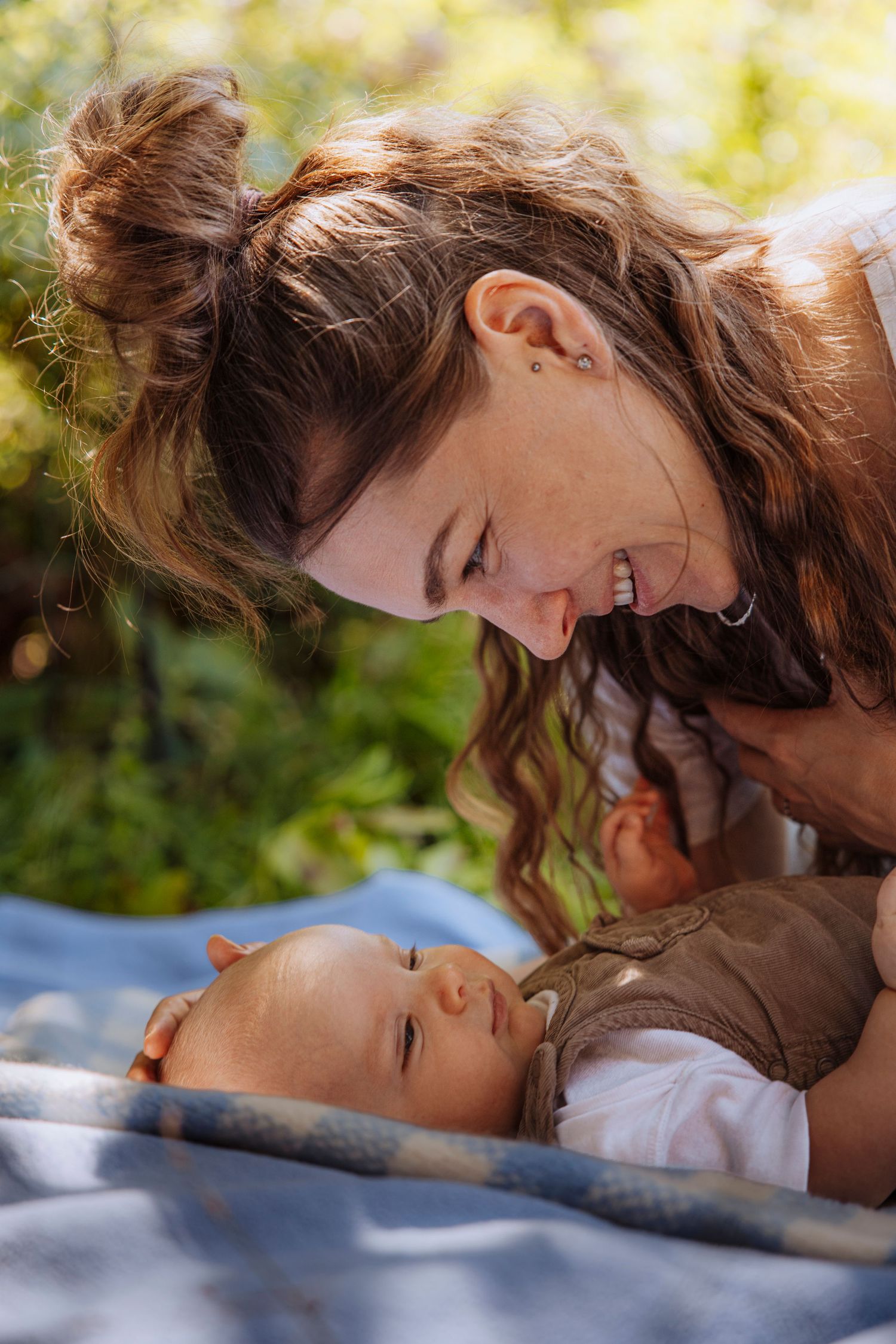 Smiling mother leaning close to her sleeping newborn baby lying on a blue blanket outdoors.