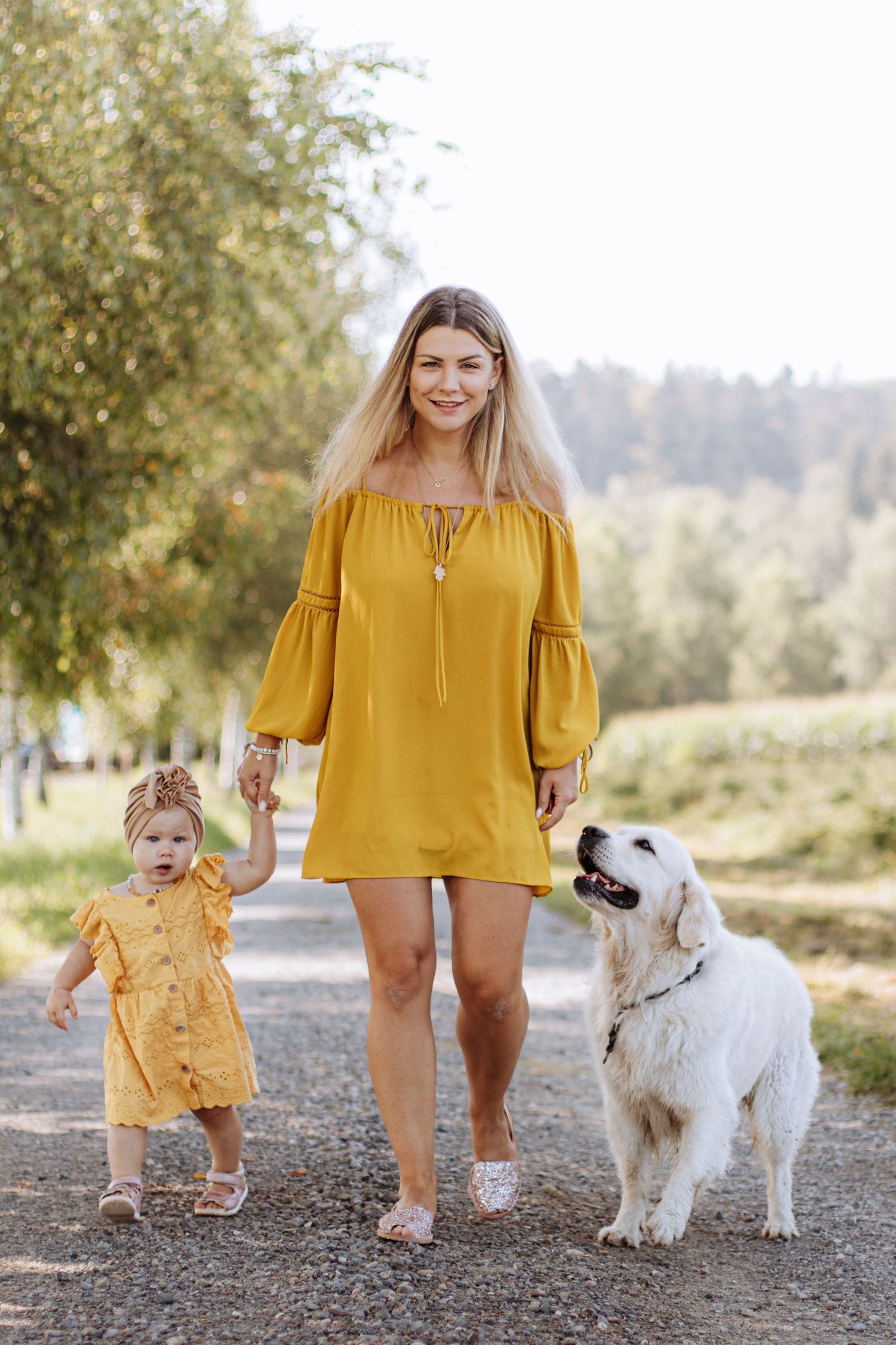 Smiling blonde mother in yellow dress walking with toddler daughter and golden retriever dog in park.