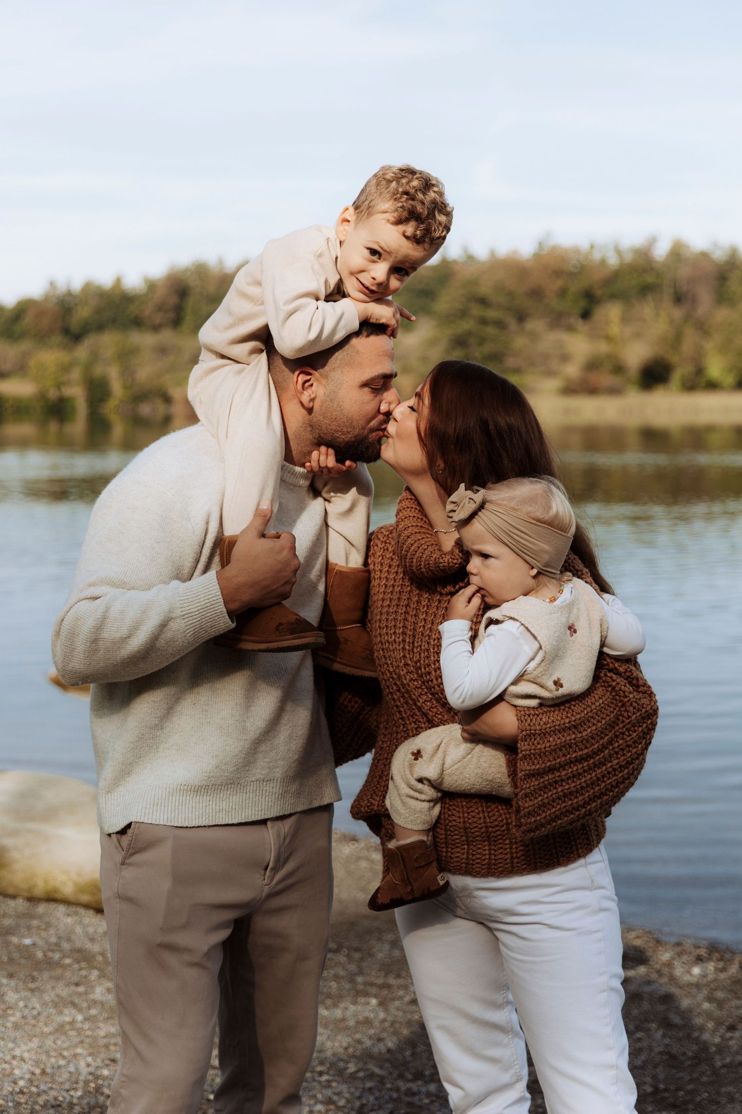 Happy family of four posing by a lake, parents kissing while holding their two young children.