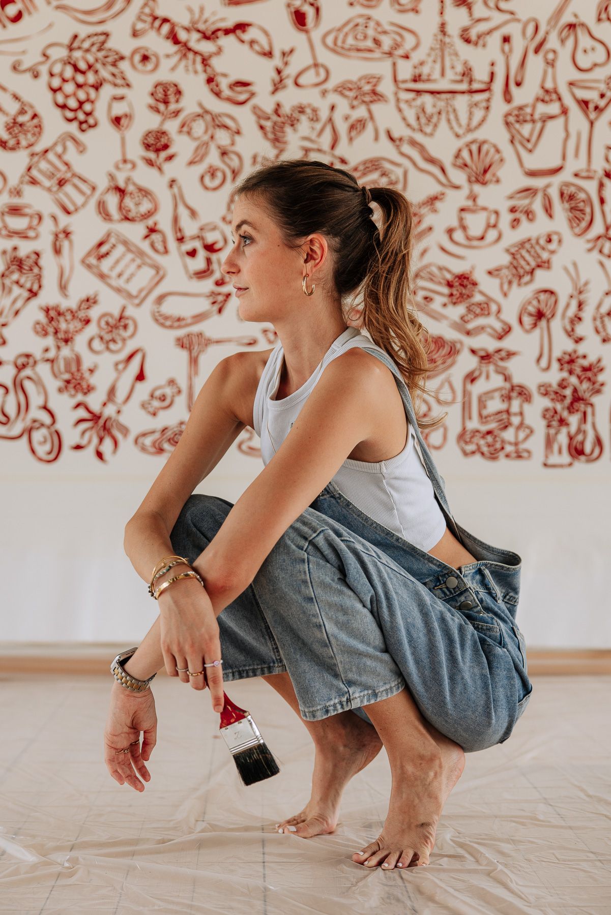 Young woman crouching barefoot in denim overalls holding a paintbrush in front of a large red illustrated mural.