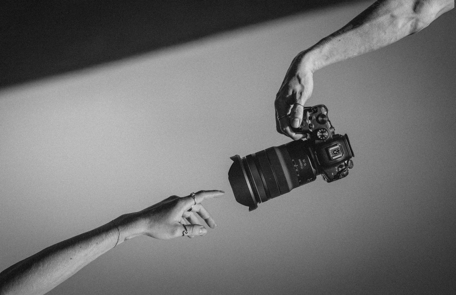 Black and white photo of two hands, one holding a DSLR camera and another pointing at the lens.