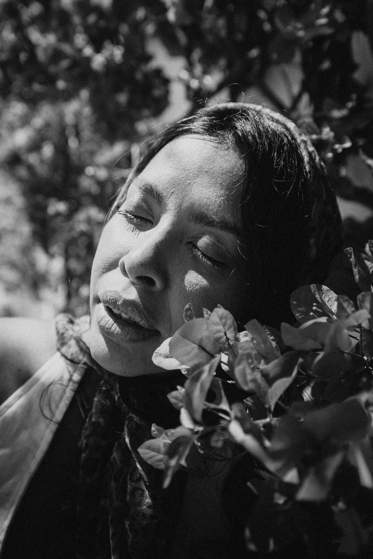 Black and white portrait of young woman with closed eyes surrounded by flowers and leaves in sunlight.