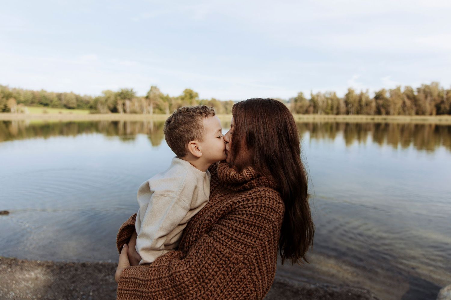 Tender moment as a young boy kisses his dark-haired mother by a serene lakeside.