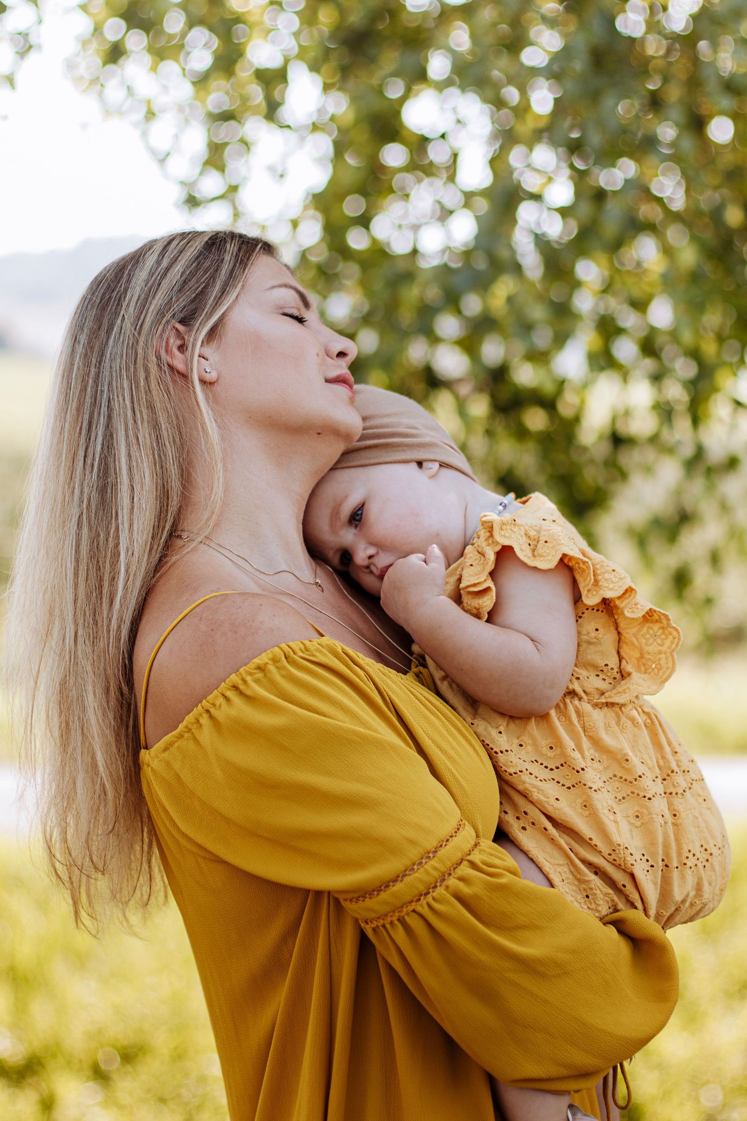 Blonde mother in yellow dress holding sleeping baby girl against her chest outdoors in sunlit park.