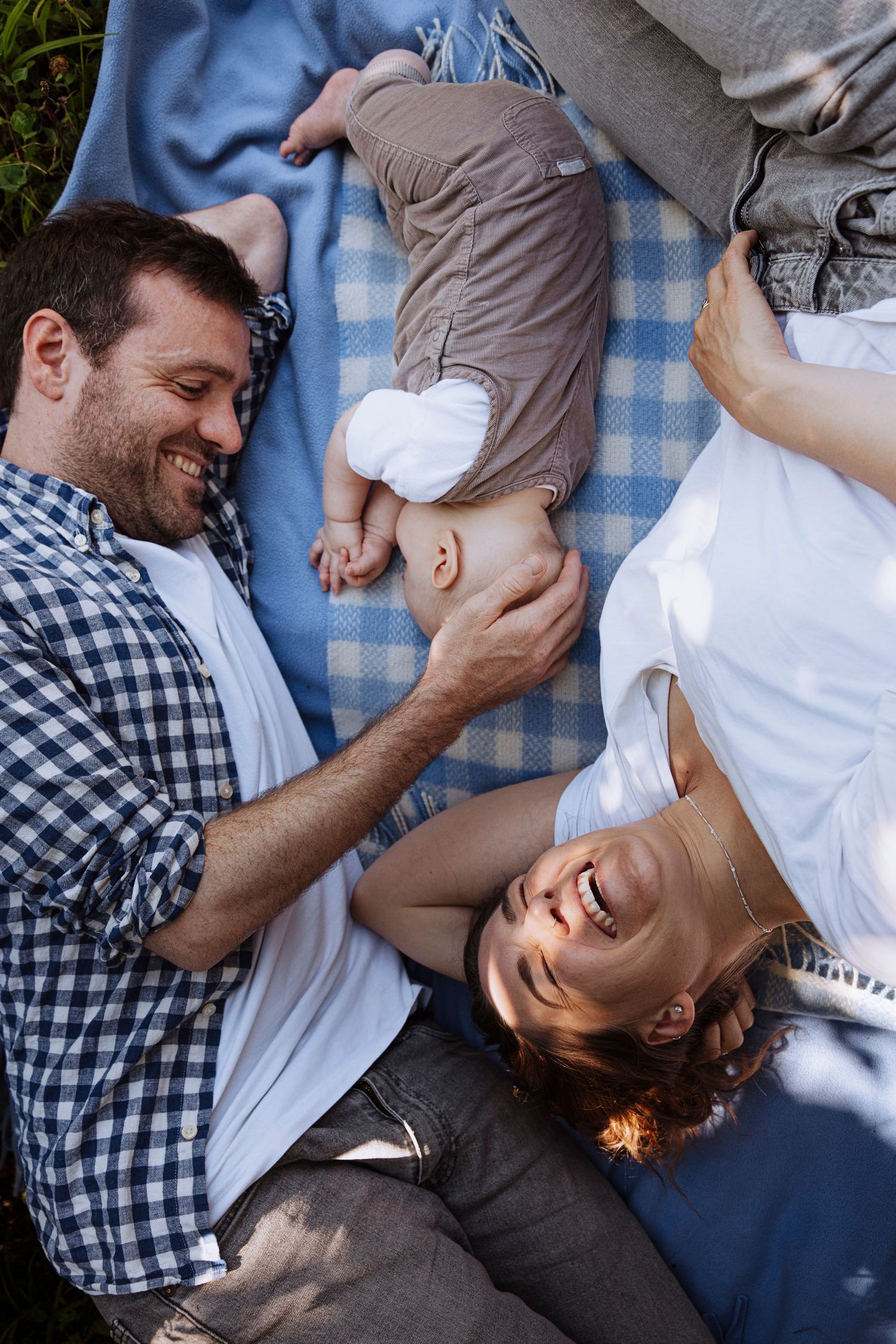 Joyful family lying on a blue checkered blanket outside, smiling and bonding with their newborn baby.