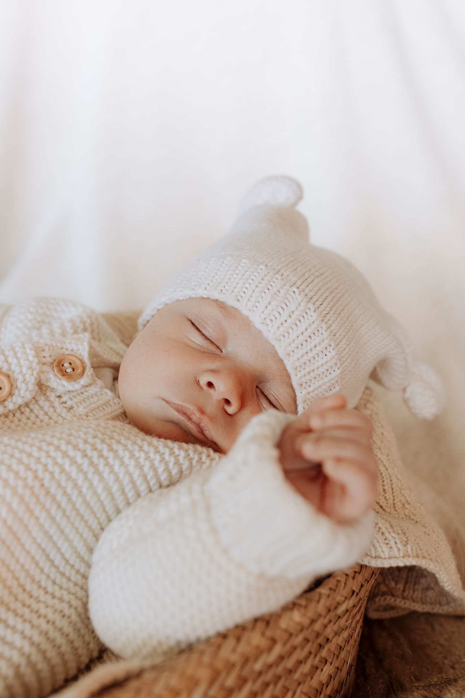 A sleeping newborn baby in a cream knit outfit and hat rests in a wicker basket.