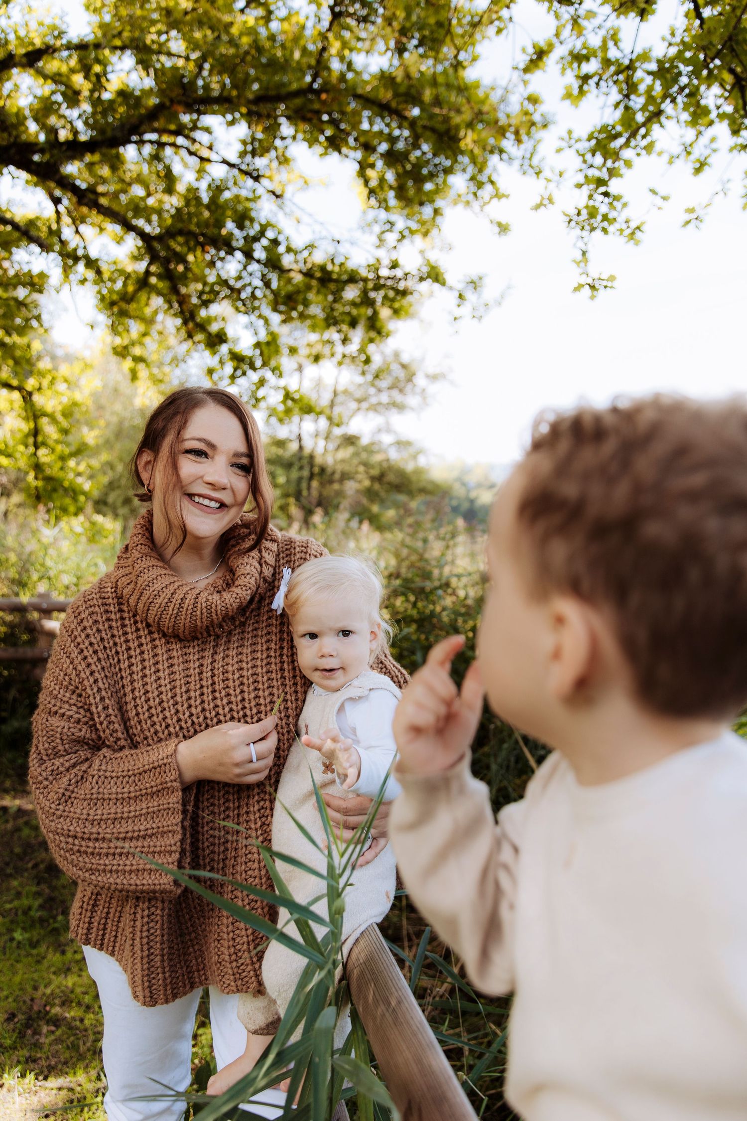 A smiling mother in a brown knit sweater holds her baby while her older child waves in a sunny park.