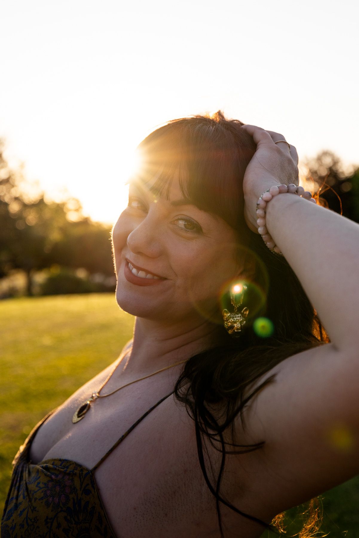 Woman smiling at sunset in a park, hand in hair, wearing a necklace and pearl bracelet, with golden backlight.