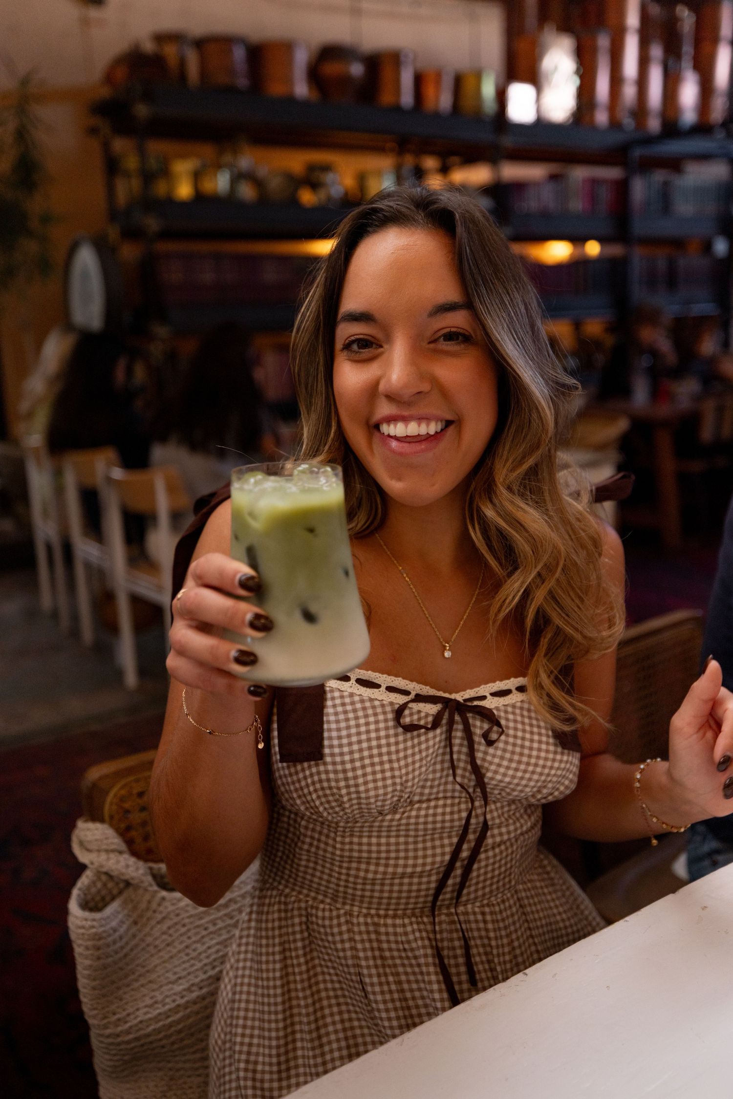 Smiling young woman holding an iced matcha latte in a cozy cafe, wearing a gingham dress.