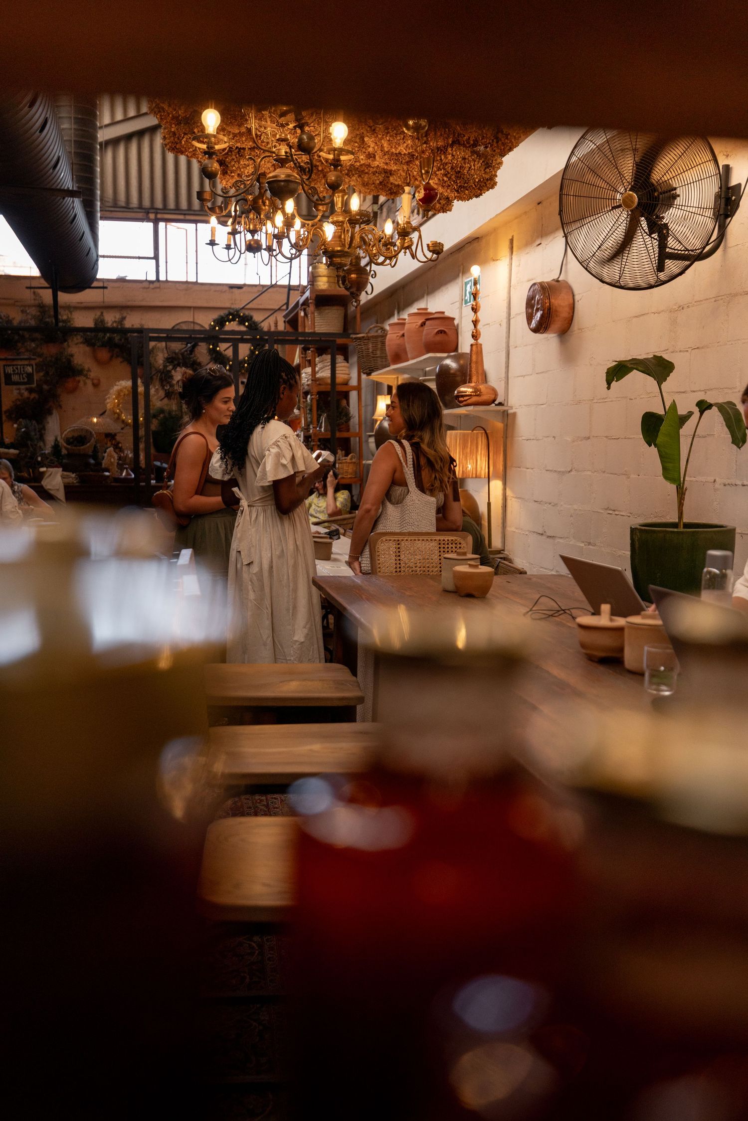 Two women in white dresses chat at a rustic cafe counter with warm Edison bulb chandelier and vintage wall fan.