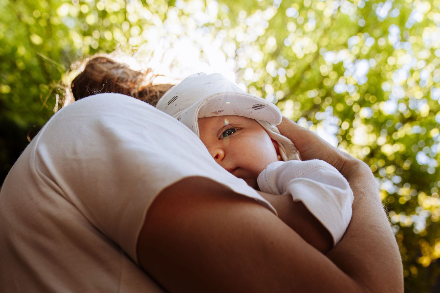Newborn baby with white hat resting peacefully on a parent's shoulder in a green garden.