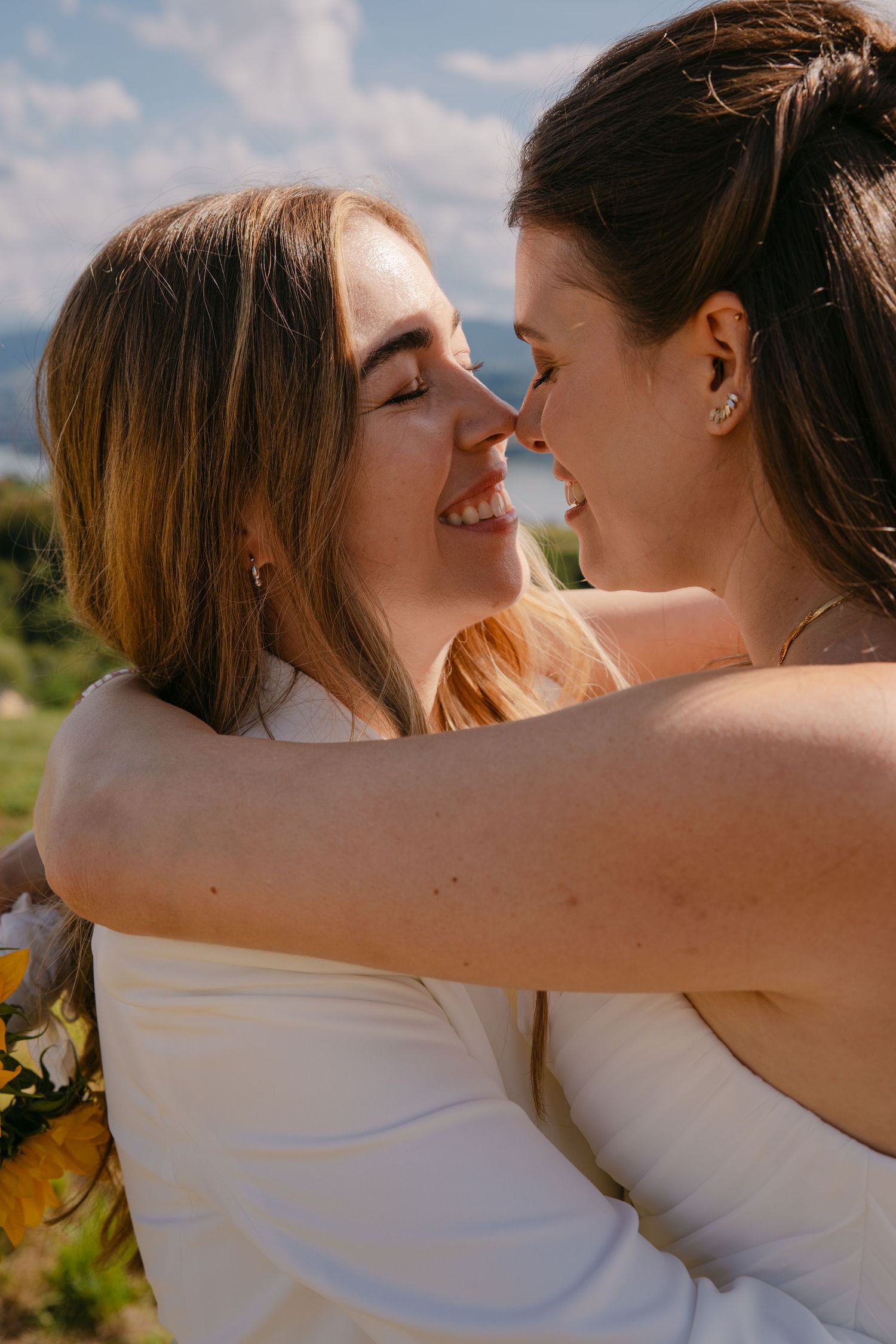 Two brides in white dresses embrace and smile at each other outdoors on a sunny wedding day.