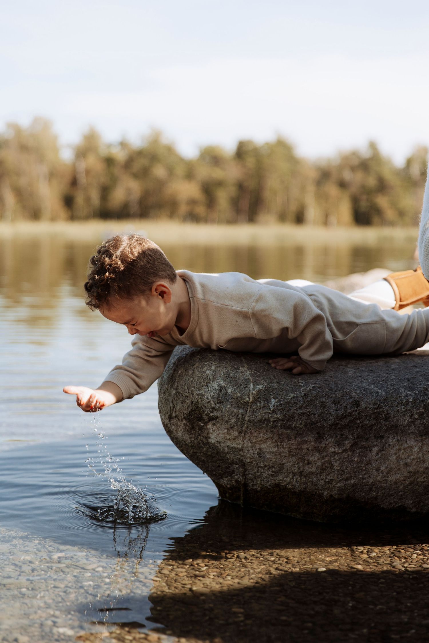 A young child lies on a rock reaching down to touch the water at a peaceful lakeside.