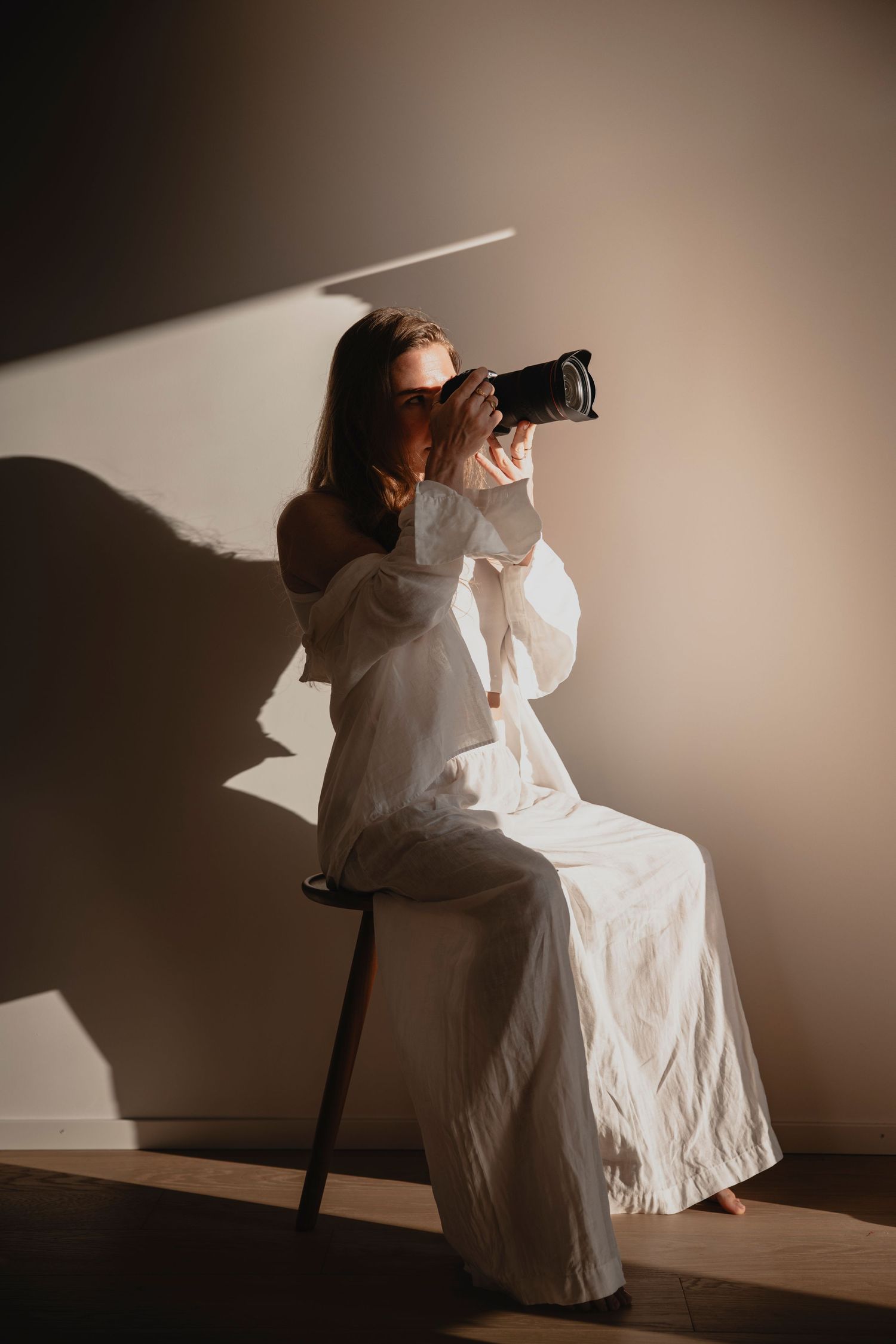 Woman in white off-shoulder outfit sitting on stool photographing with DSLR camera indoors.