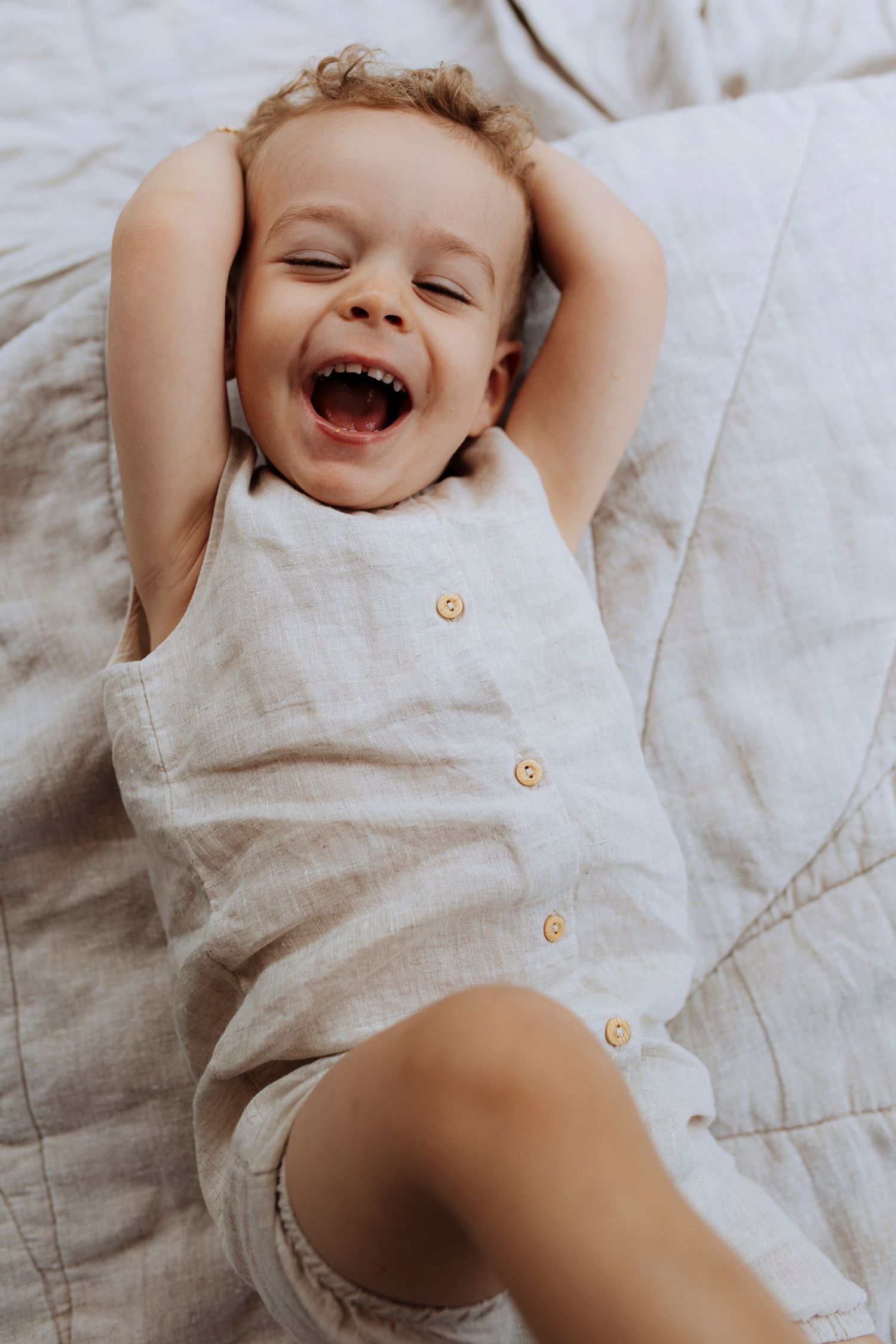 A happy toddler in a linen romper laughs joyfully while lying on a soft bed.