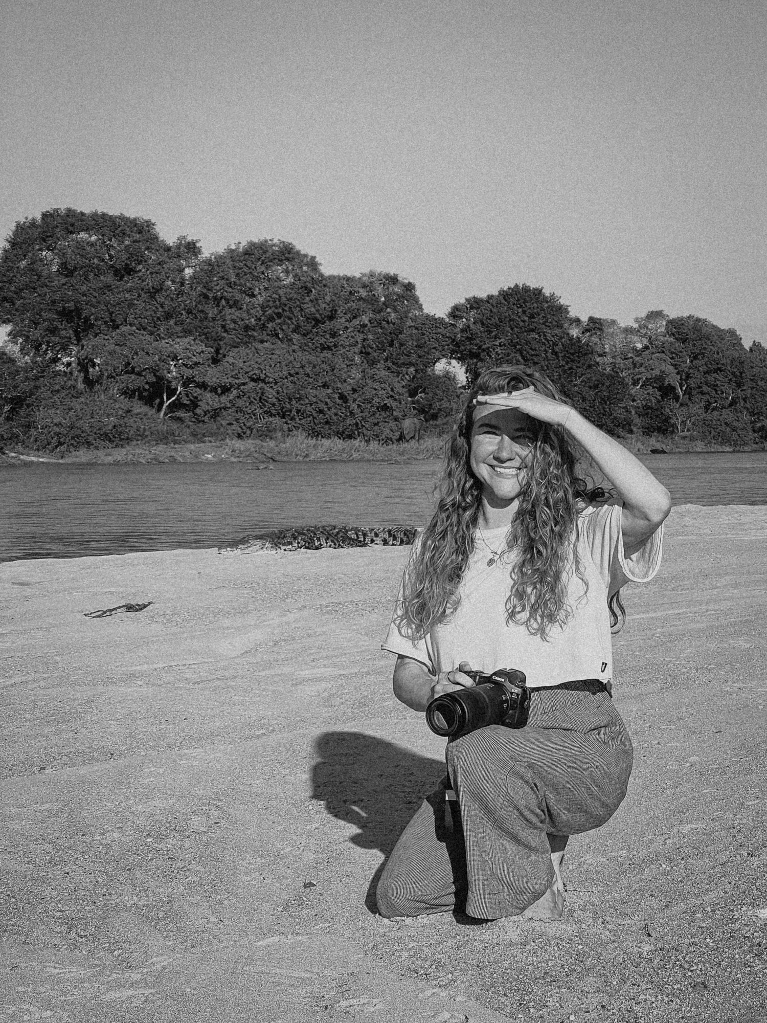 Woman crouching on sandy riverbank holding camera, shielding eyes from sun, with trees and water in black and white photo.