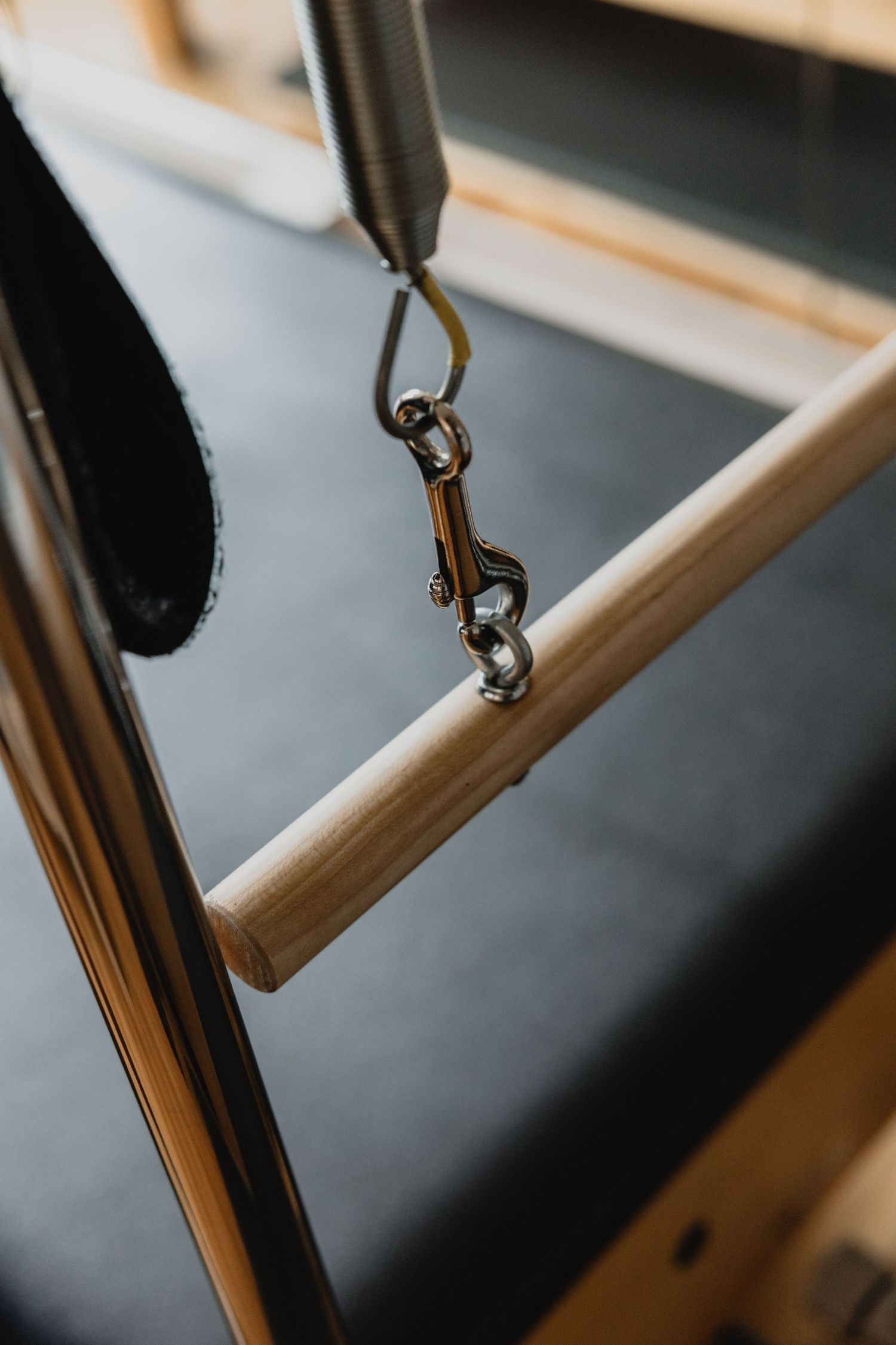 Close-up of a metal carabiner clip attached to a wooden bar on a Pilates reformer machine in a studio.