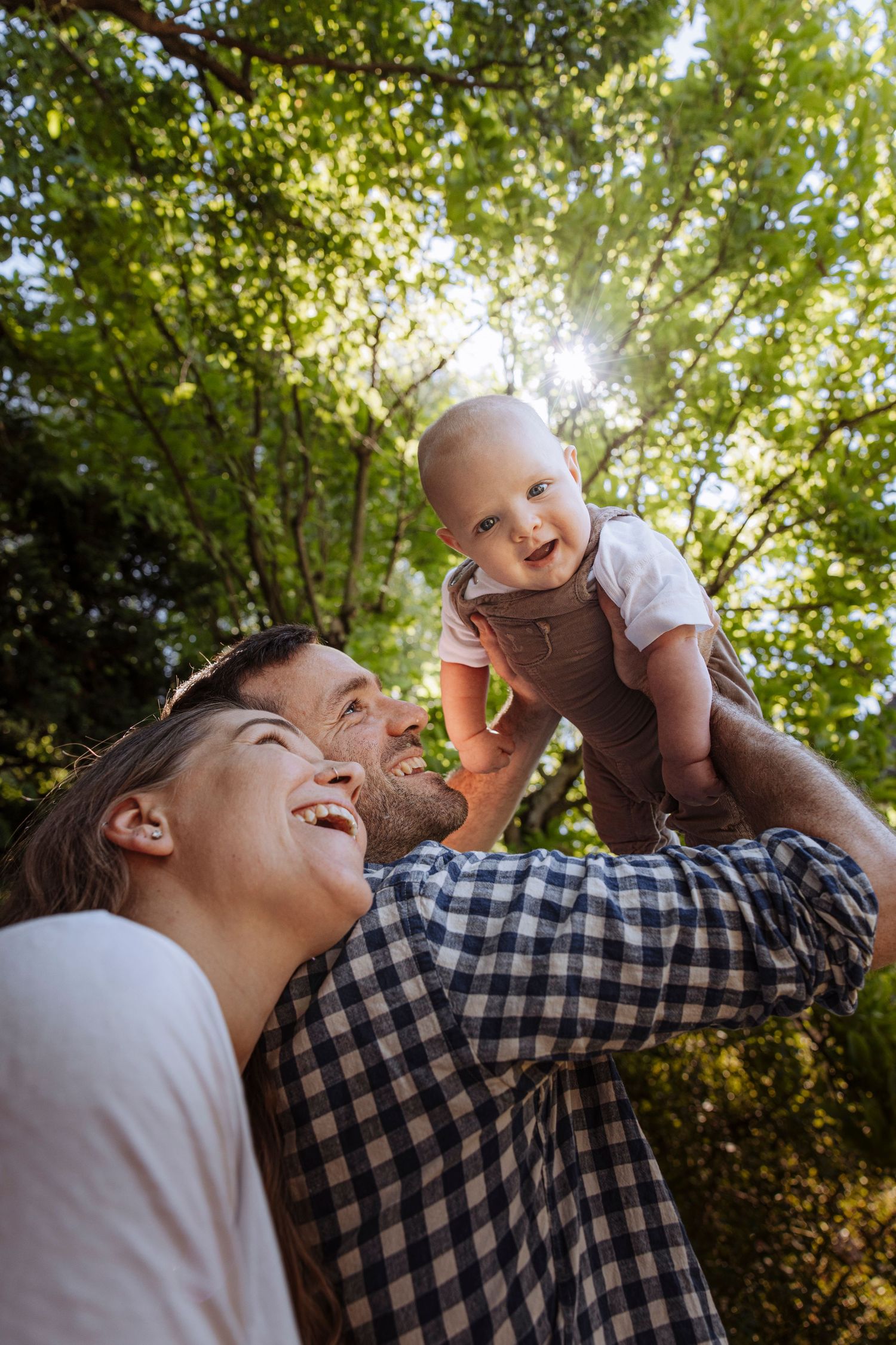 Happy young parents laughing while father lifts smiling baby up in a sunny green park.