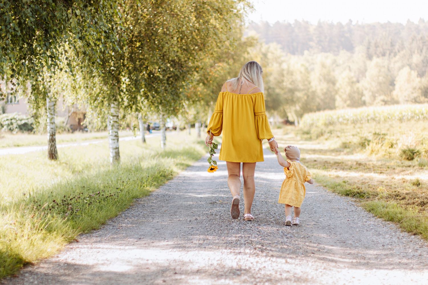 Mother and toddler daughter in matching yellow outfits walking hand in hand down a sunny tree-lined path.