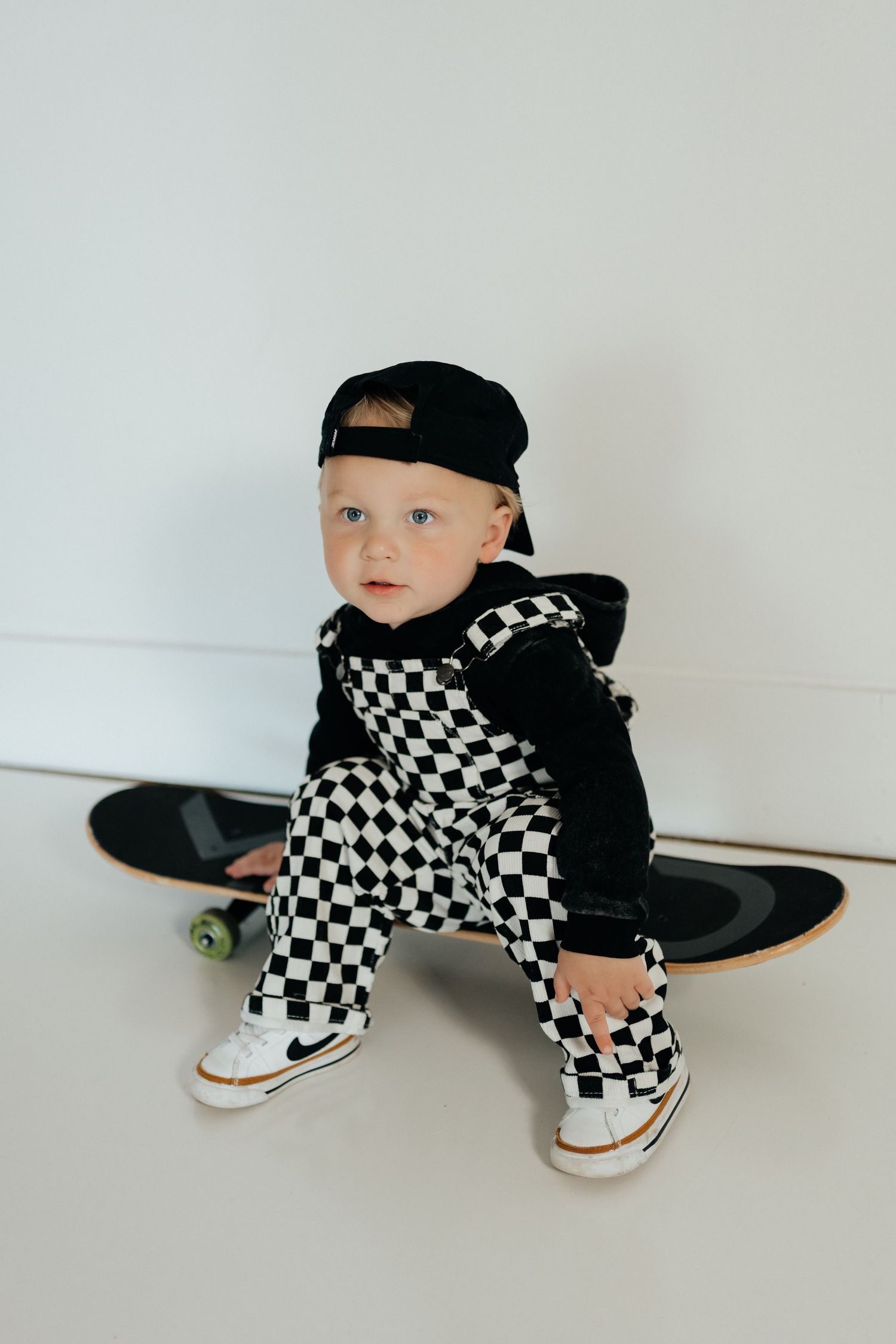 Toddler boy in checkered overalls, black hoodie, cap, and Nike sneakers sitting on a skateboard against a white wall.