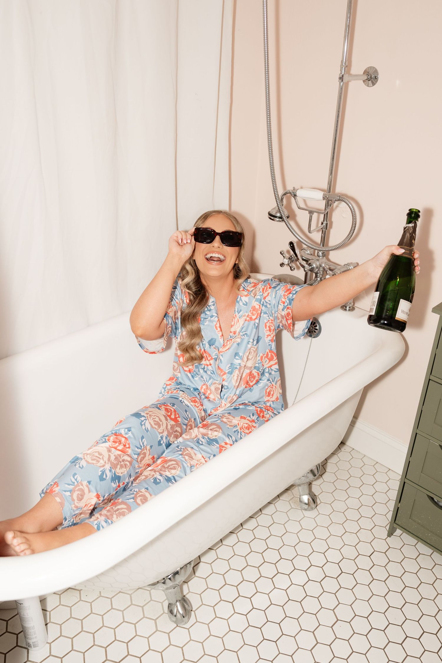 Woman in floral jumpsuit and sunglasses relaxing in clawfoot bathtub holding champagne bottle.