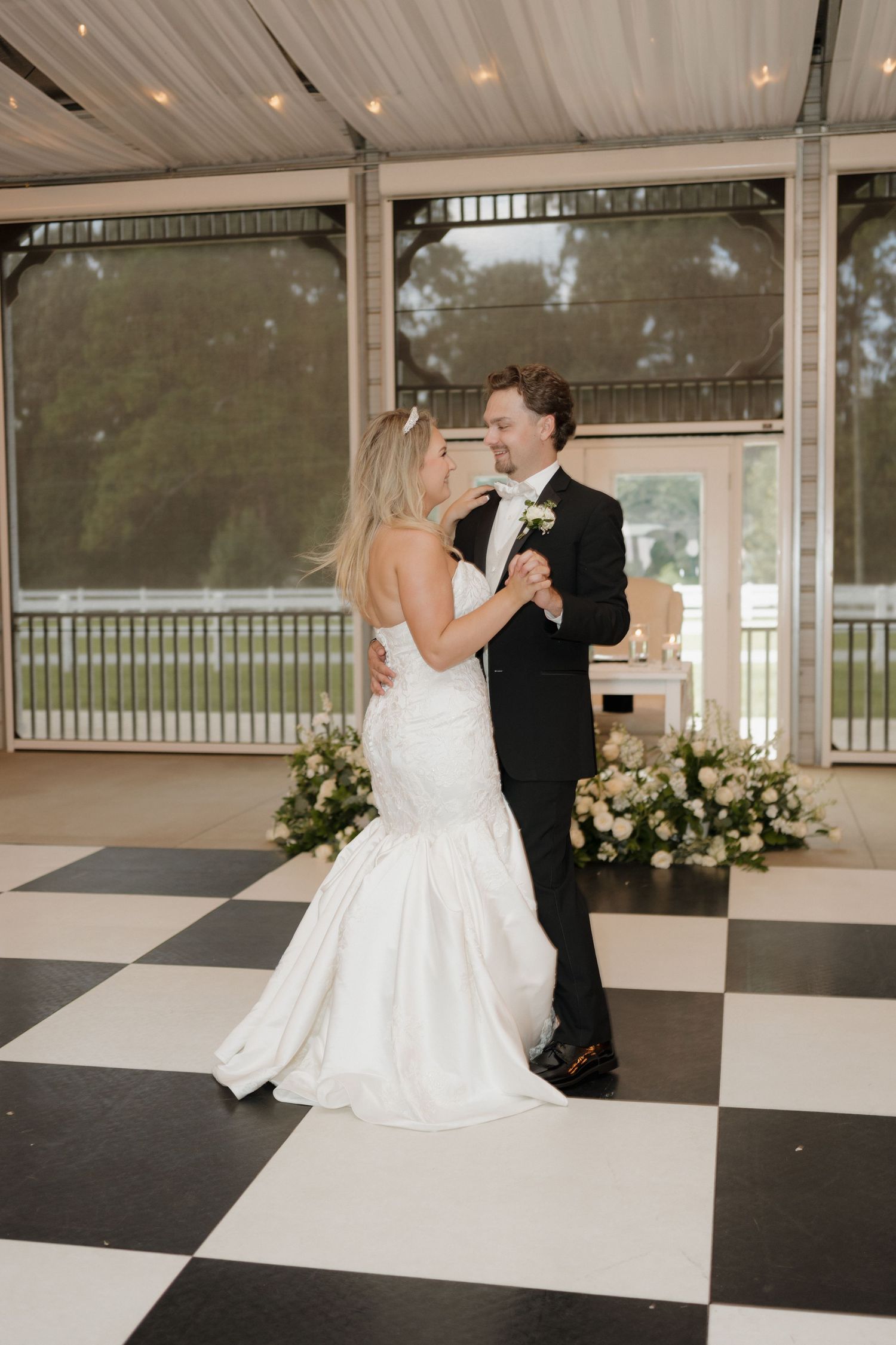 Newlywed couple shares their first dance on a black and white checkered floor in an elegant covered venue.