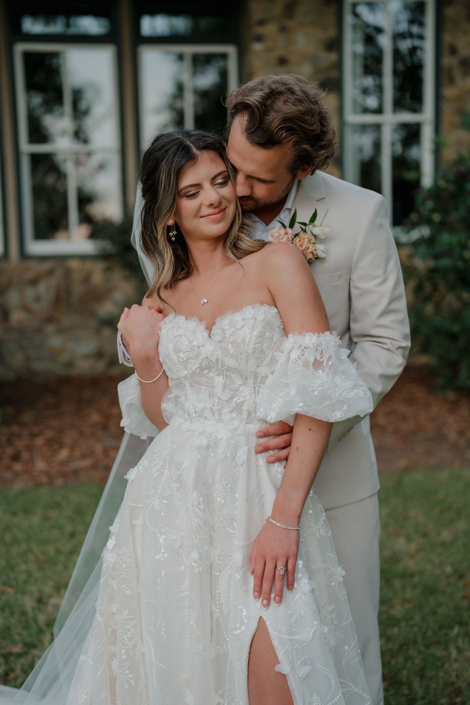 Intimate wedding portrait of couple embracing in front of stone building with bride in detailed lace gown.
