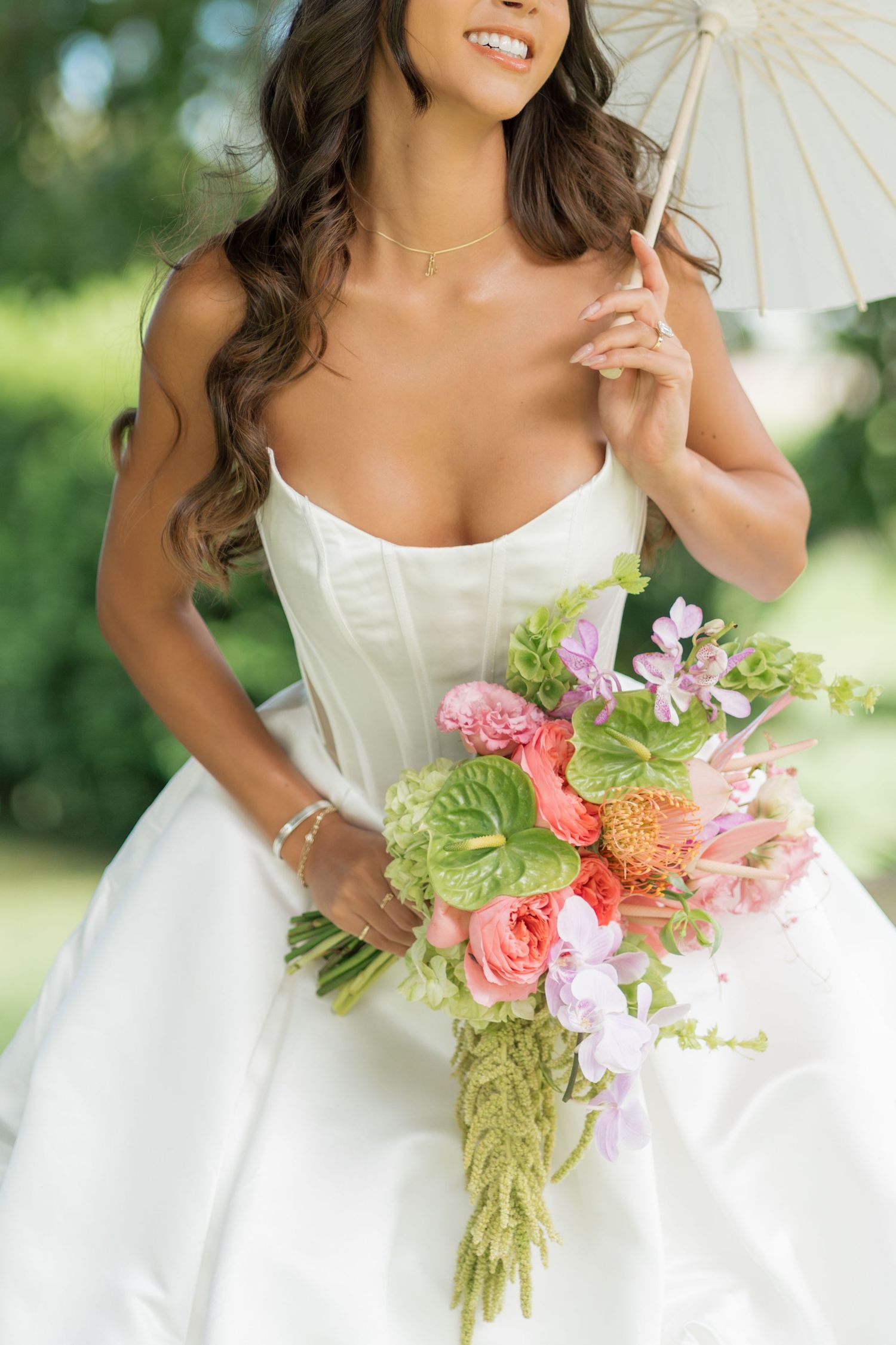 Bride in white gown holding colorful floral bouquet and parasol, smiling outdoors on her wedding day.