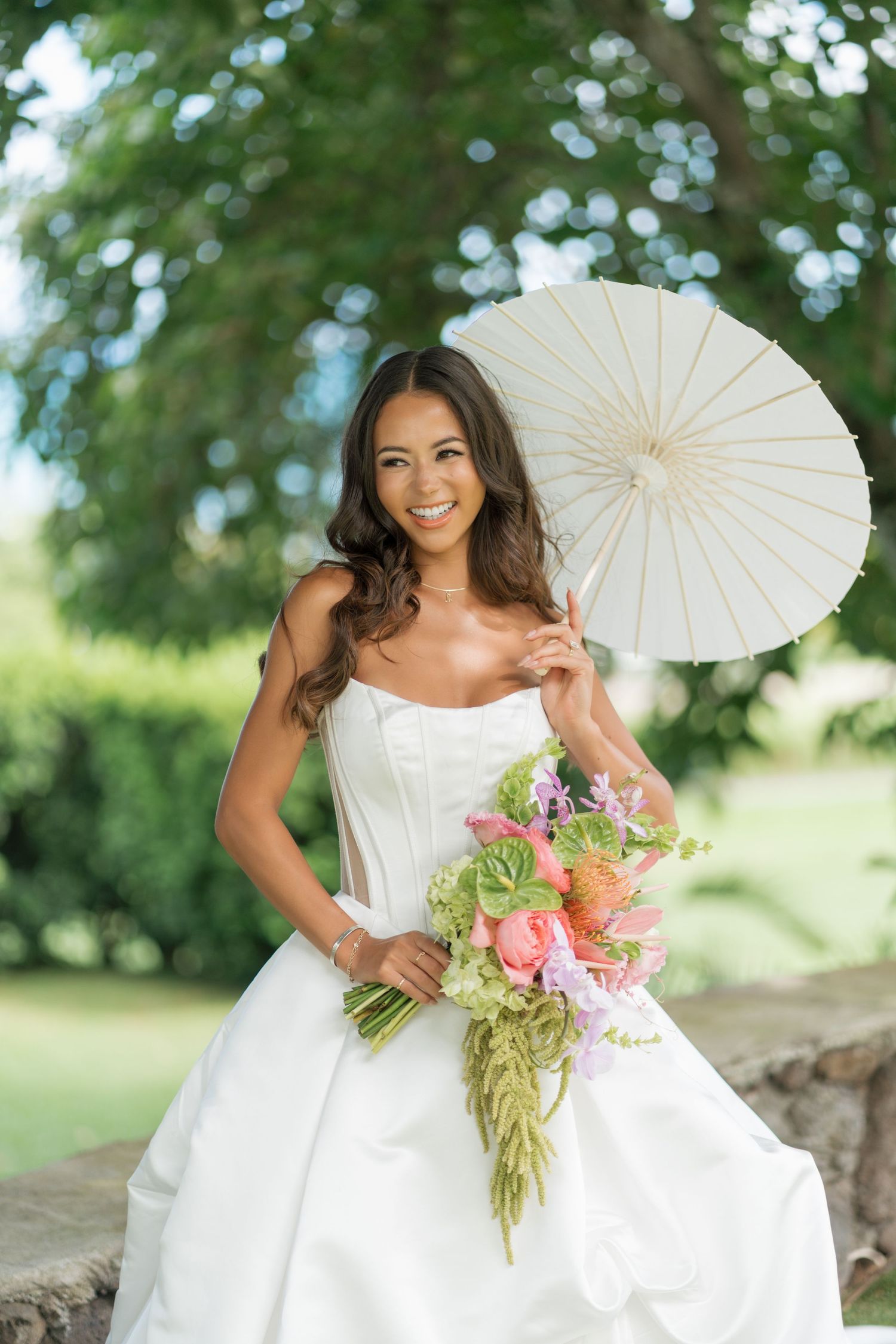 Smiling bride in white strapless gown holds pink floral bouquet and white parasol umbrella outdoors in lush green garden.