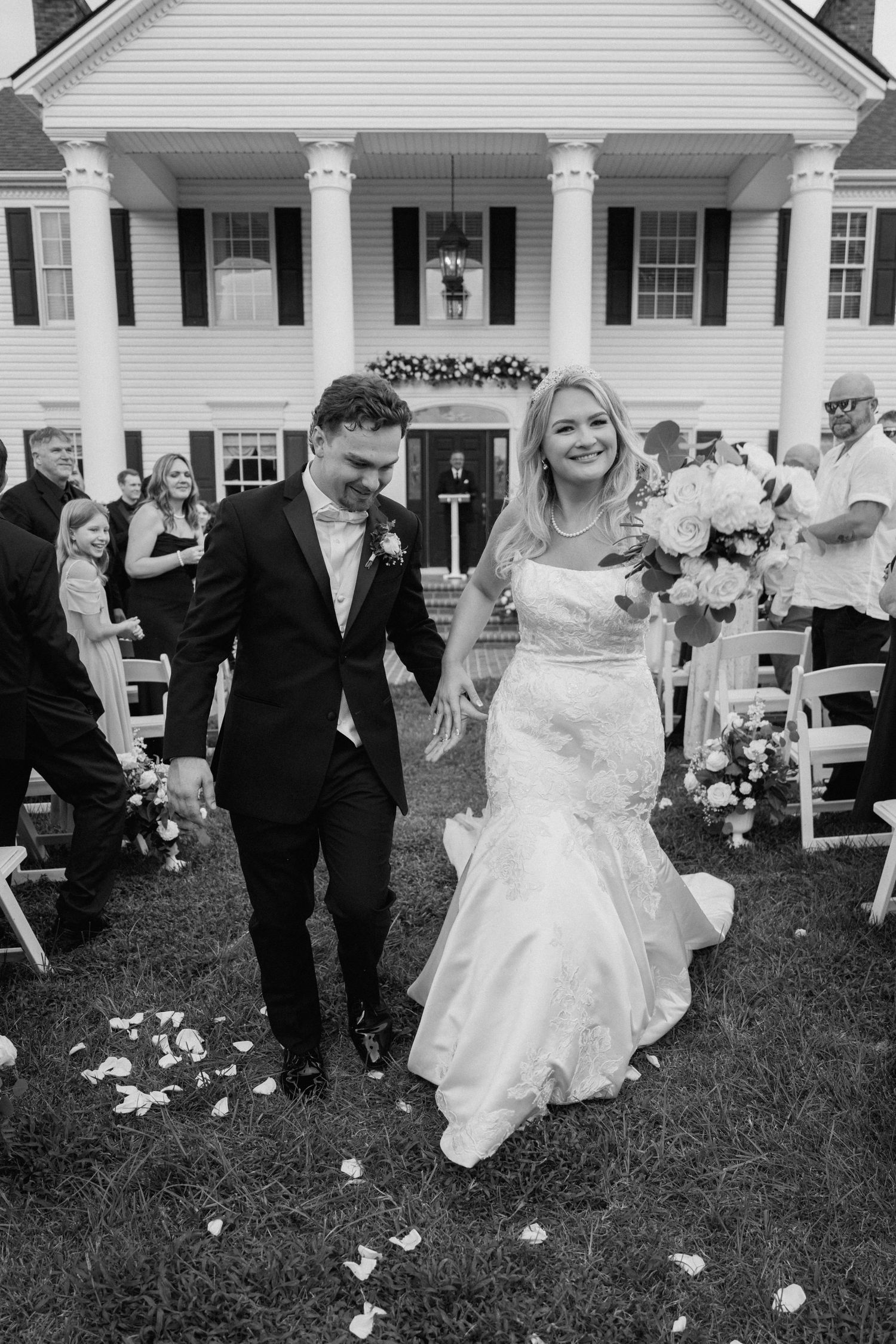 Outdoor wedding celebration on lawn of white colonial house with scattered flower petals along the aisle.