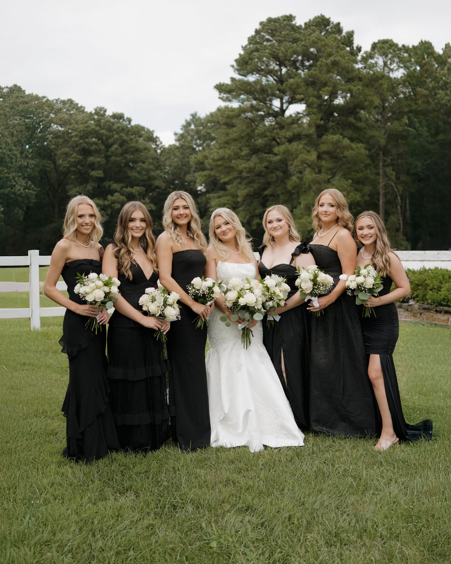 A bride in white poses with bridesmaids wearing elegant black dresses on a green lawn with tall trees in the background.