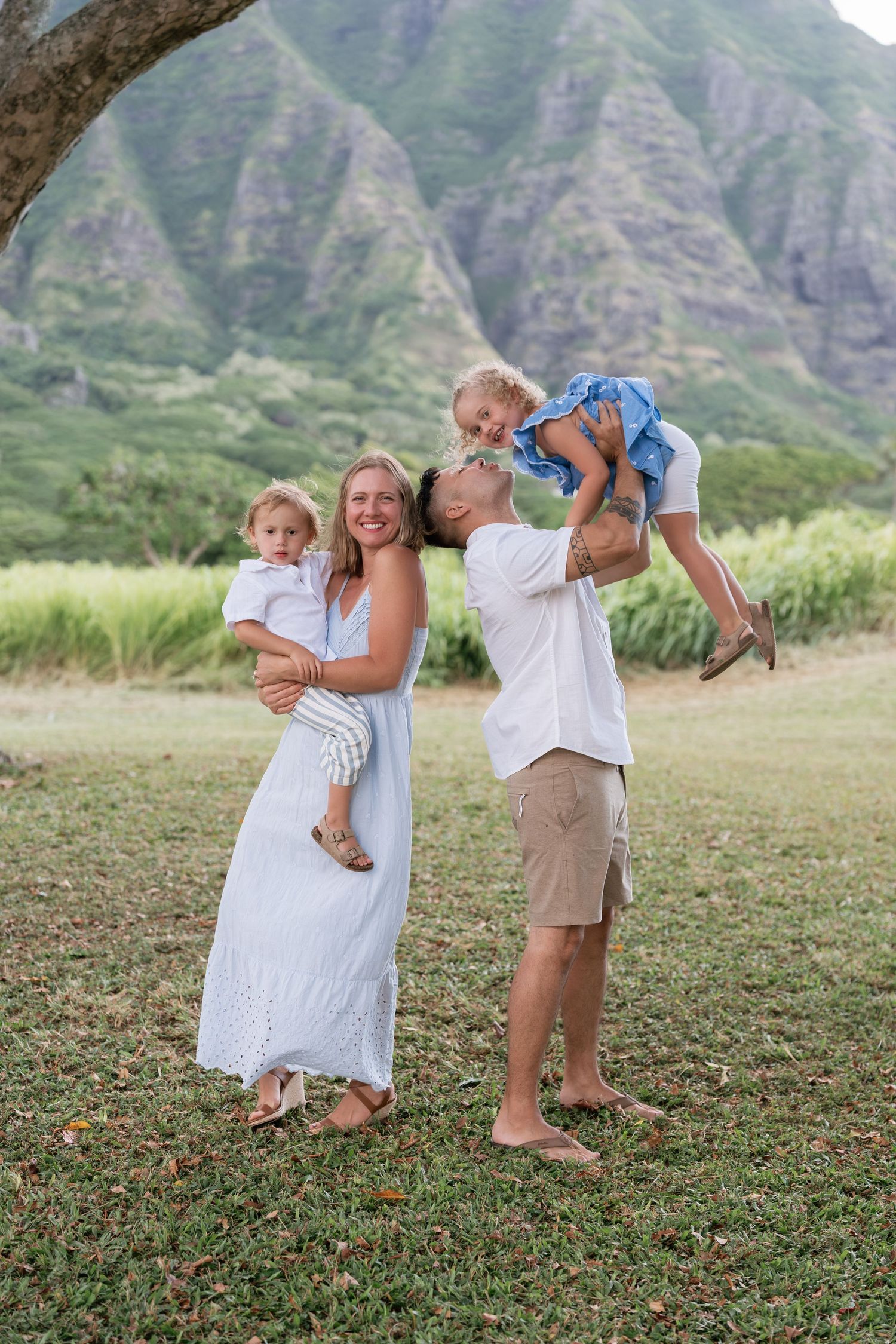 Family of four posing outdoors with lush green mountains, parents holding two young children, wearing white and neutral tones.