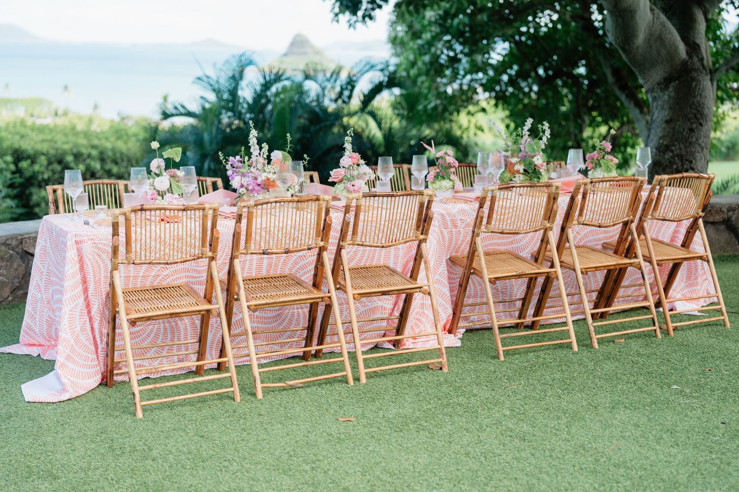 Outdoor wedding reception table with pink floral linen, rattan chairs, and tropical floral centerpieces on green lawn.