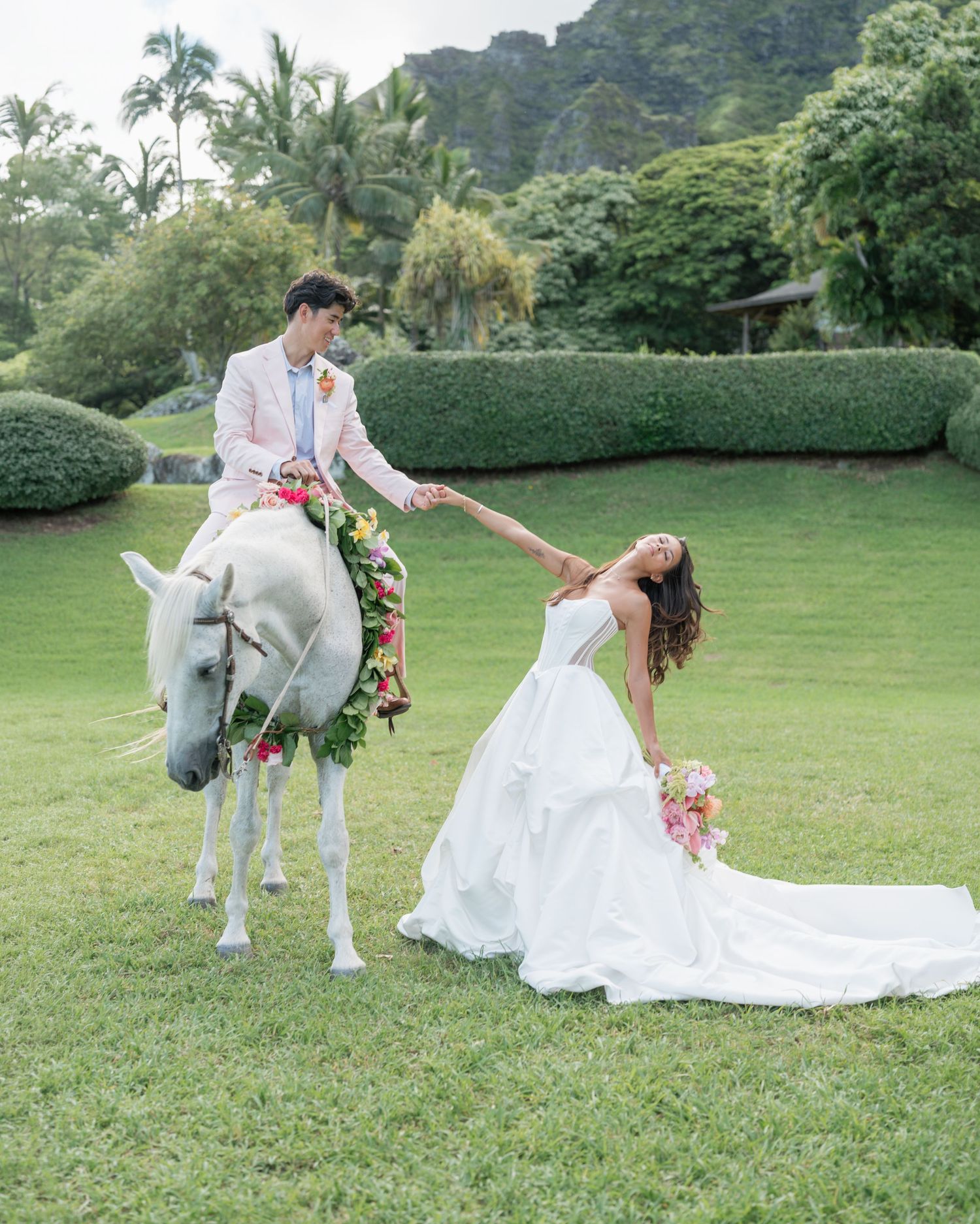 Bride in white gown holds groom's hand as he sits on a flower-adorned white horse at a tropical garden wedding.