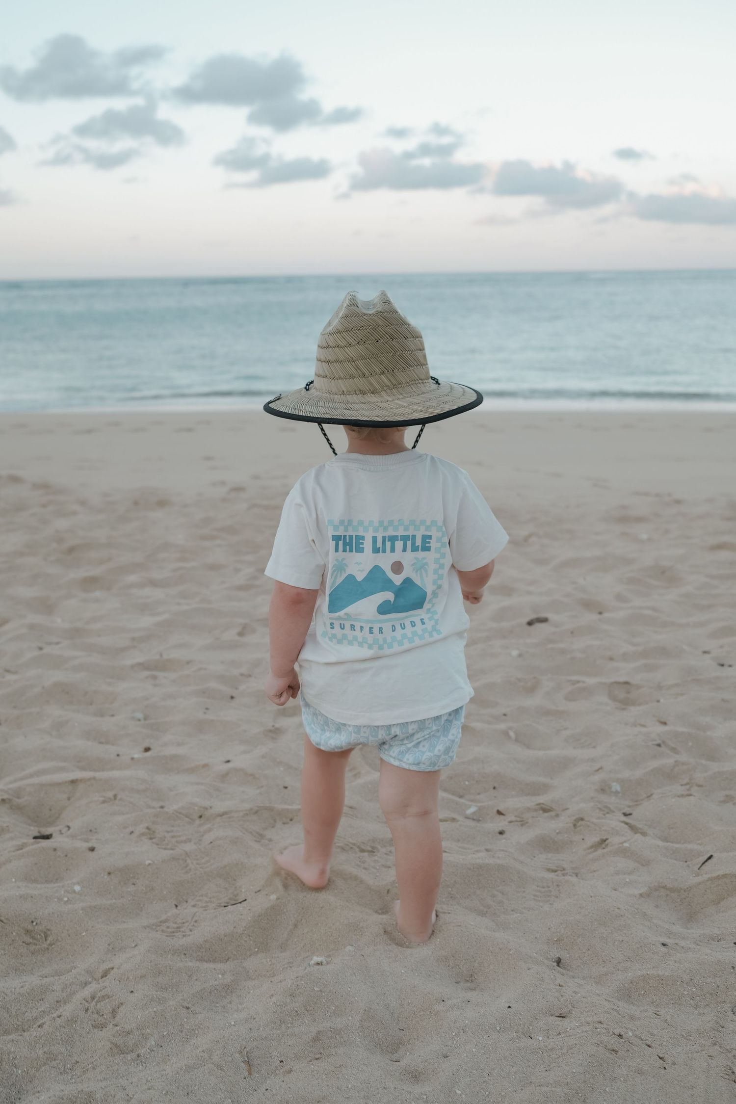 Toddler in straw hat and white graphic tee stands barefoot on sandy beach facing calm ocean at sunset.