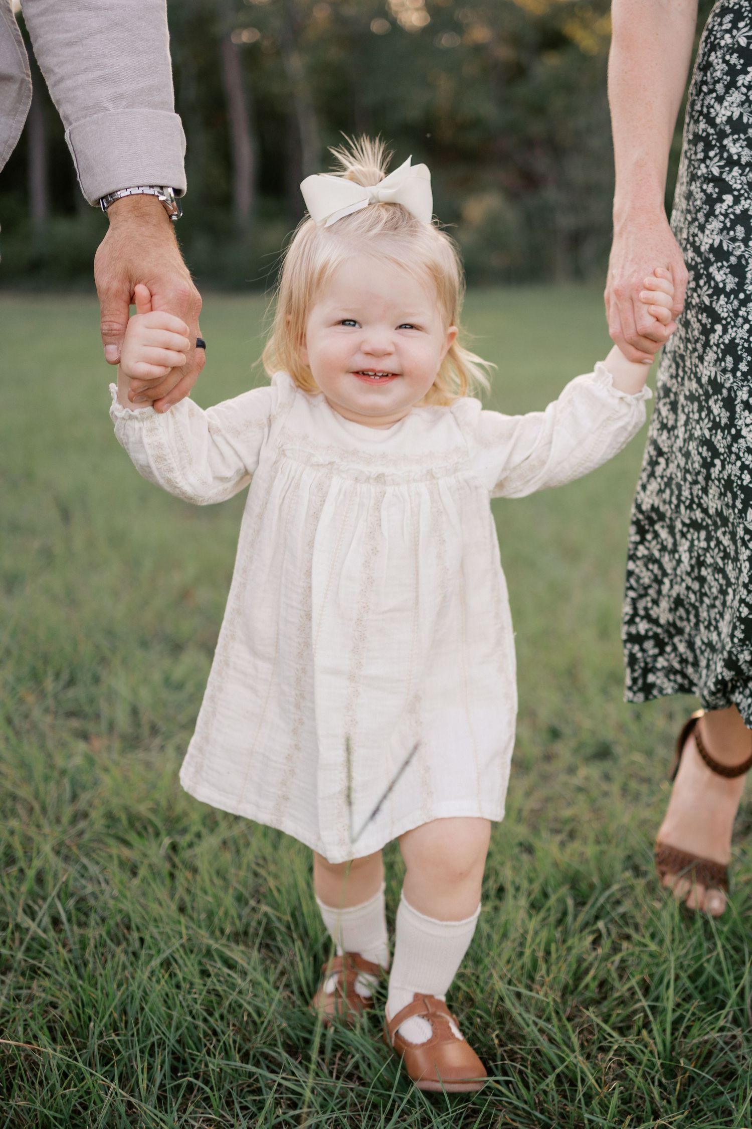 A small child in a white dress holds hands with adults while standing on grass outdoors.