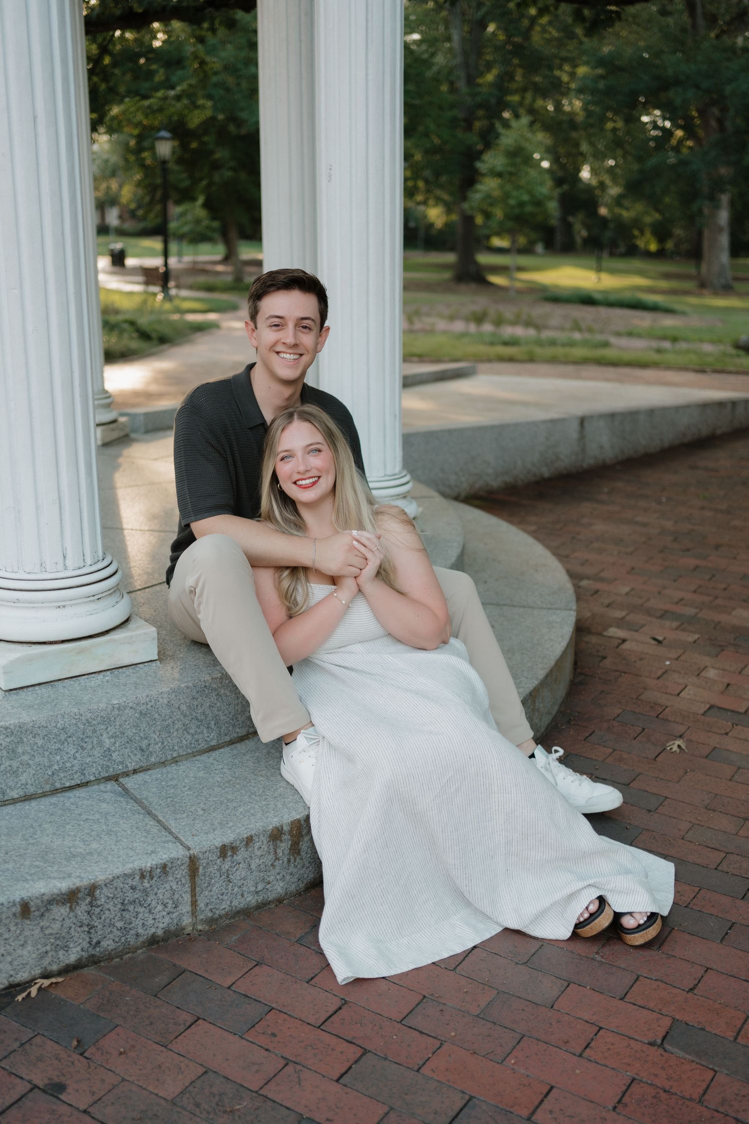 A couple sits together on stone steps by white columns on a brick pathway in an outdoor setting.