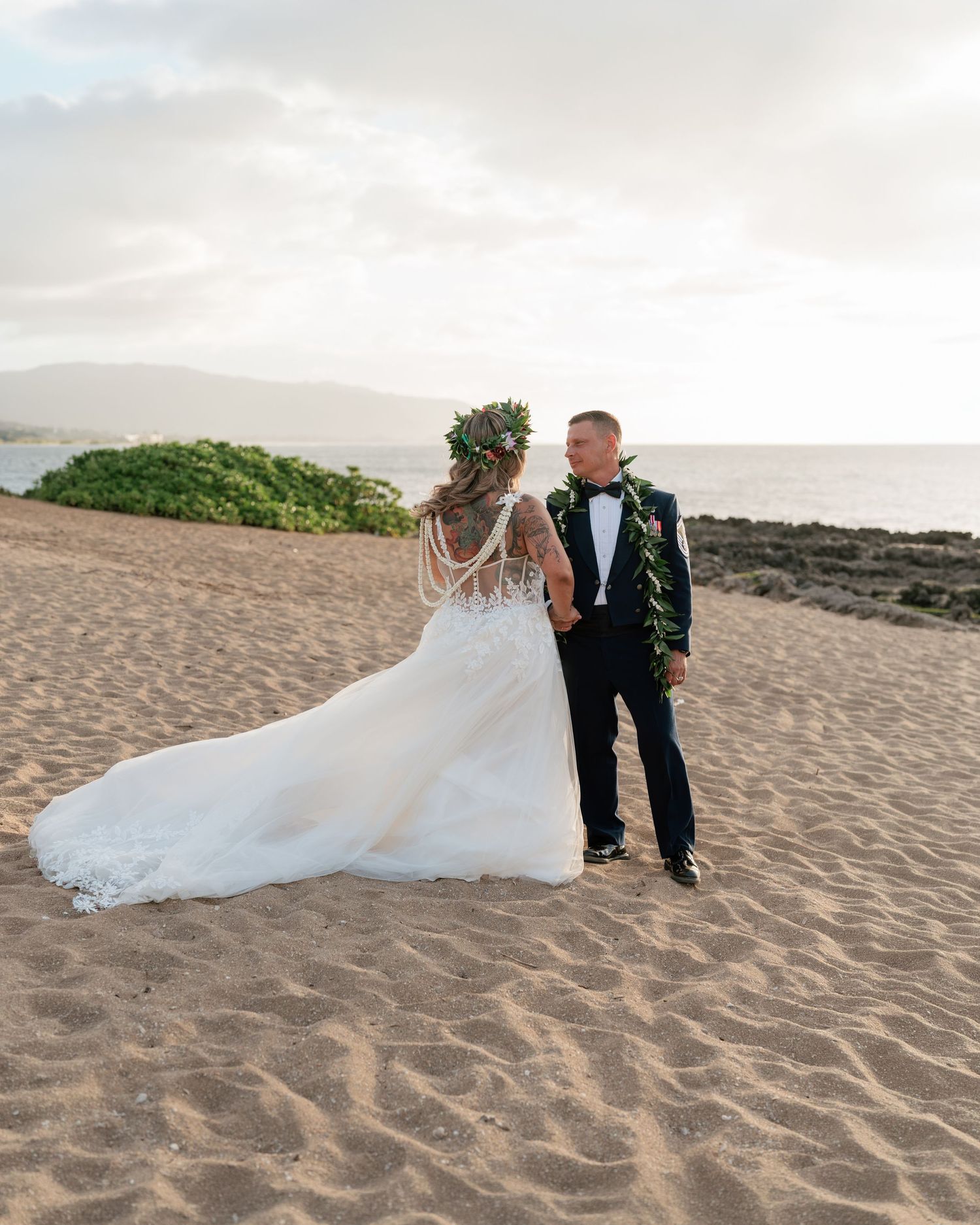Bride and groom stand on sandy beach at sunset, she wears floral crown and long train gown, he wears dark tuxedo.