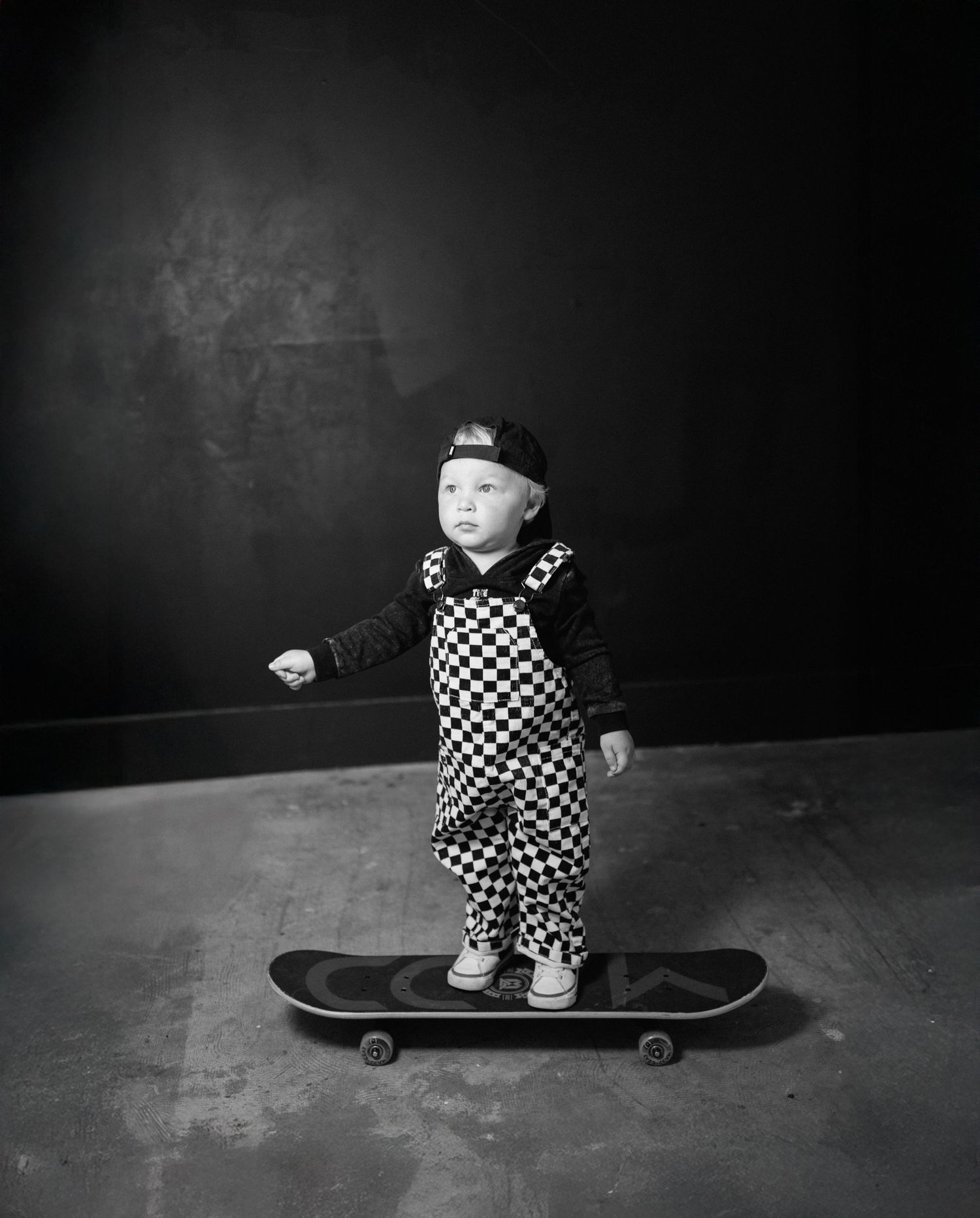 Black and white photo of a toddler in checkered overalls and cap standing on a skateboard.