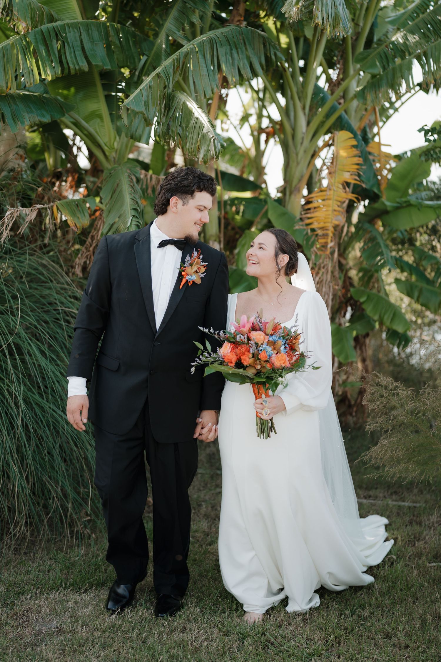 A couple shares a romantic moment in their wedding attire against a tropical backdrop with palm trees and lush greenery.