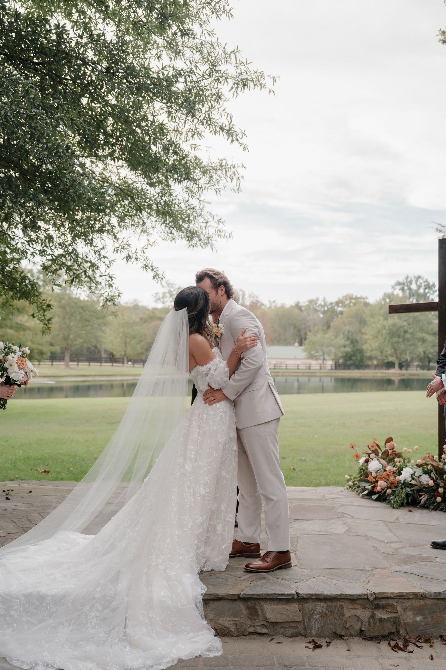 A romantic wedding ceremony kiss on a stone platform overlooking a serene lake with flowing white veil and grey suit.