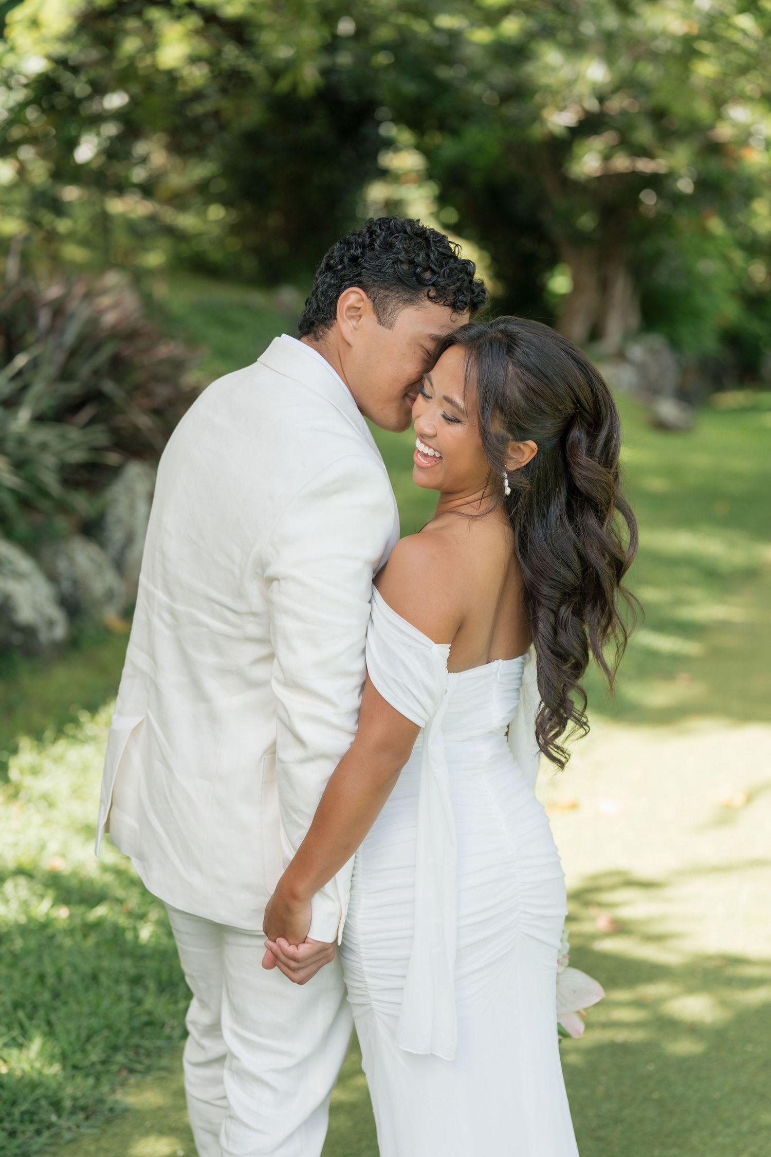 A couple in white wedding attire embrace and smile in a lush garden setting, the bride in an off-shoulder gown.