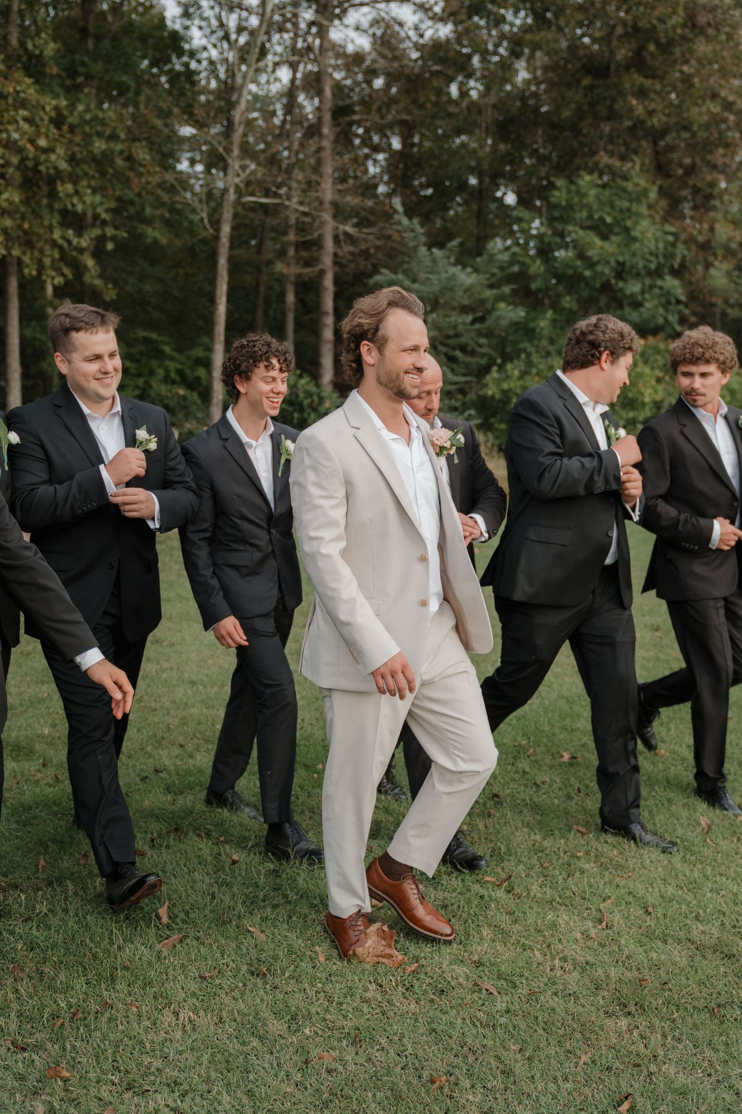 Groomsmen in black suits and groom in white perform fun jumping poses on grassy lawn during wedding photoshoot.