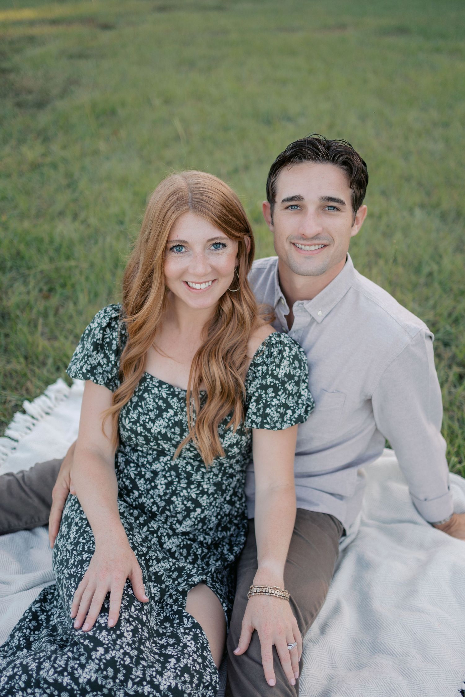 A couple sits together on a white blanket in a grassy field during an outdoor photo session.