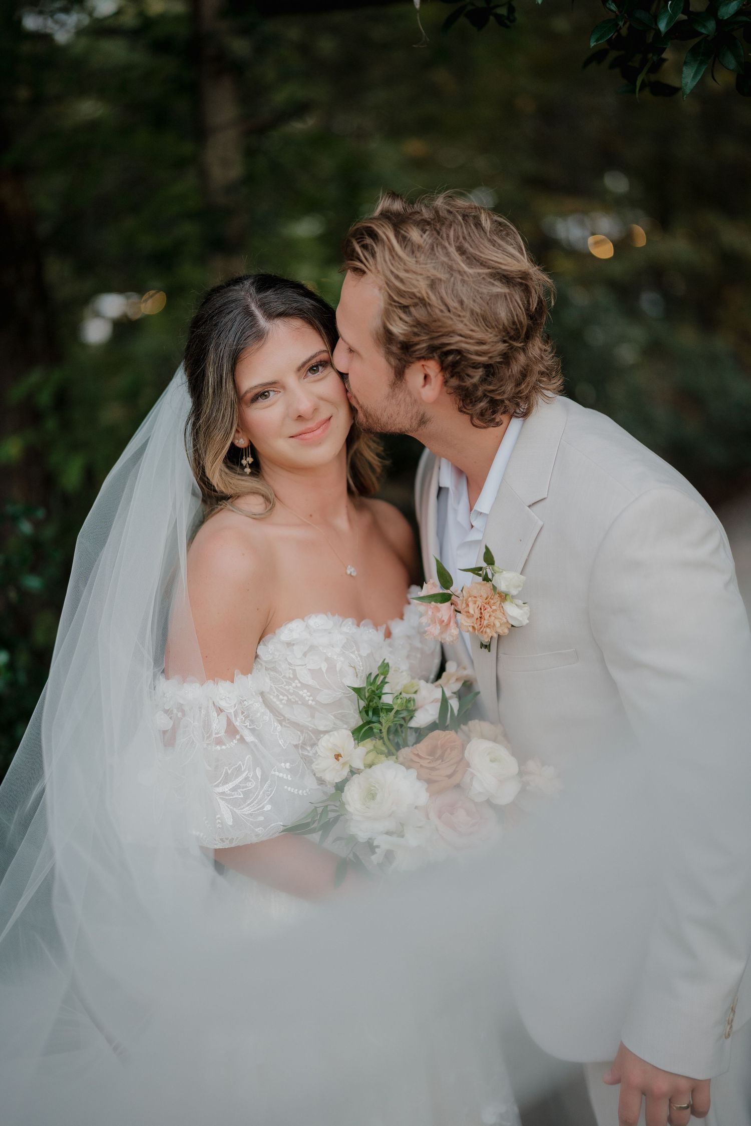 Romantic wedding couple embraces in an intimate moment outdoors with flowing white veil and light colored attire.