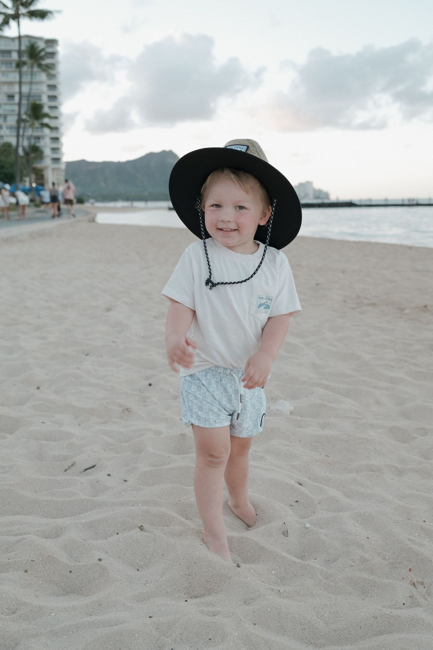 Smiling toddler wearing a black wide-brim hat and white tee plays on sandy Waikiki Beach with Diamond Head in background.