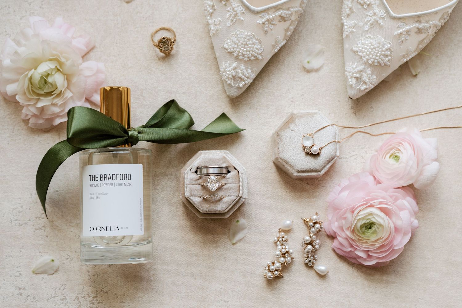 Elegant bridal flat lay featuring The Bradford perfume, diamond rings, pearl earrings, lace heels, and pink ranunculus flowers.