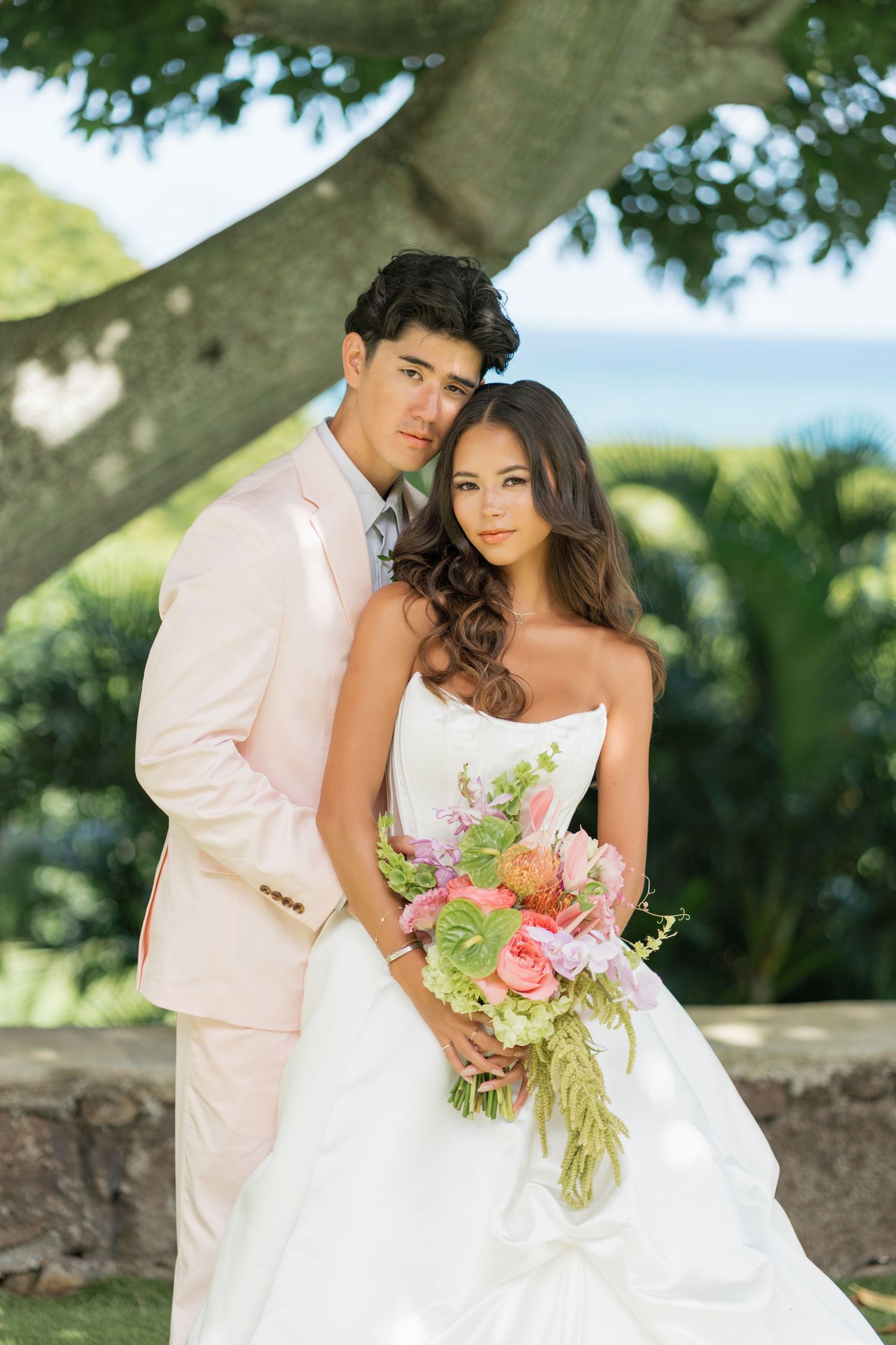 Bride and groom pose under a tree, she holds a pink bouquet in a white gown, he wears a blush suit.