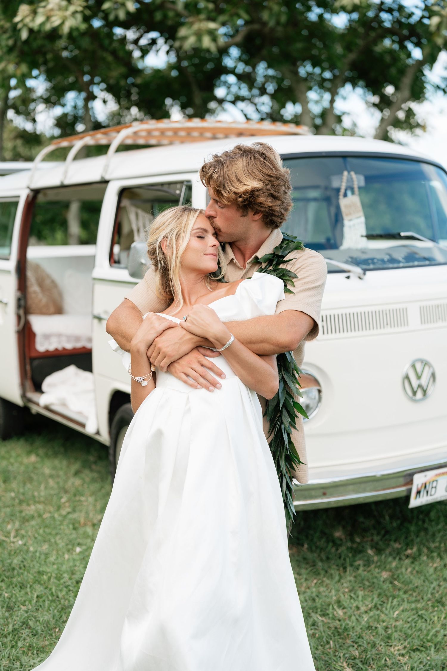 Couple in wedding attire embracing in front of a vintage white VW bus on a lush green lawn.