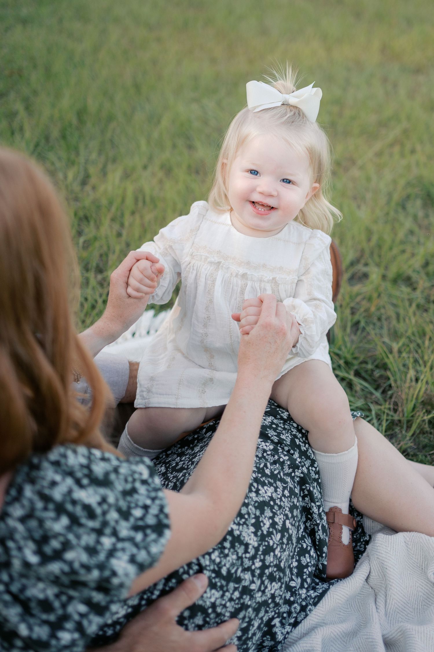 A baby in a white dress smiles while sitting on grass with someone in a floral dress.